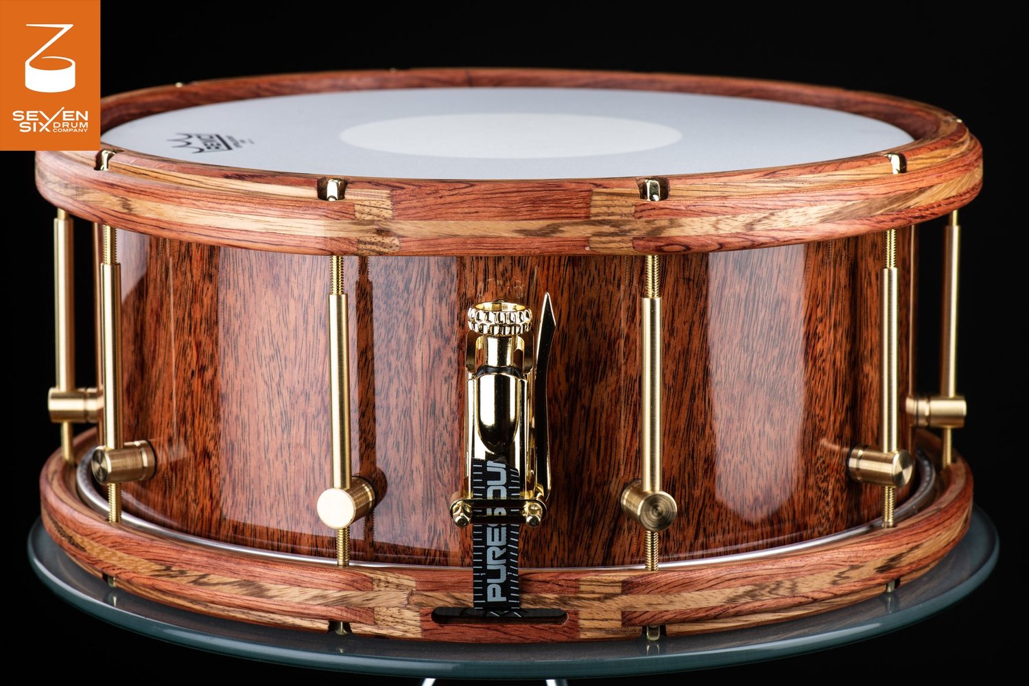 Glossy wood snare drum with segmented wooden hoops, brass tube lugs, and a polished gold throw-off.