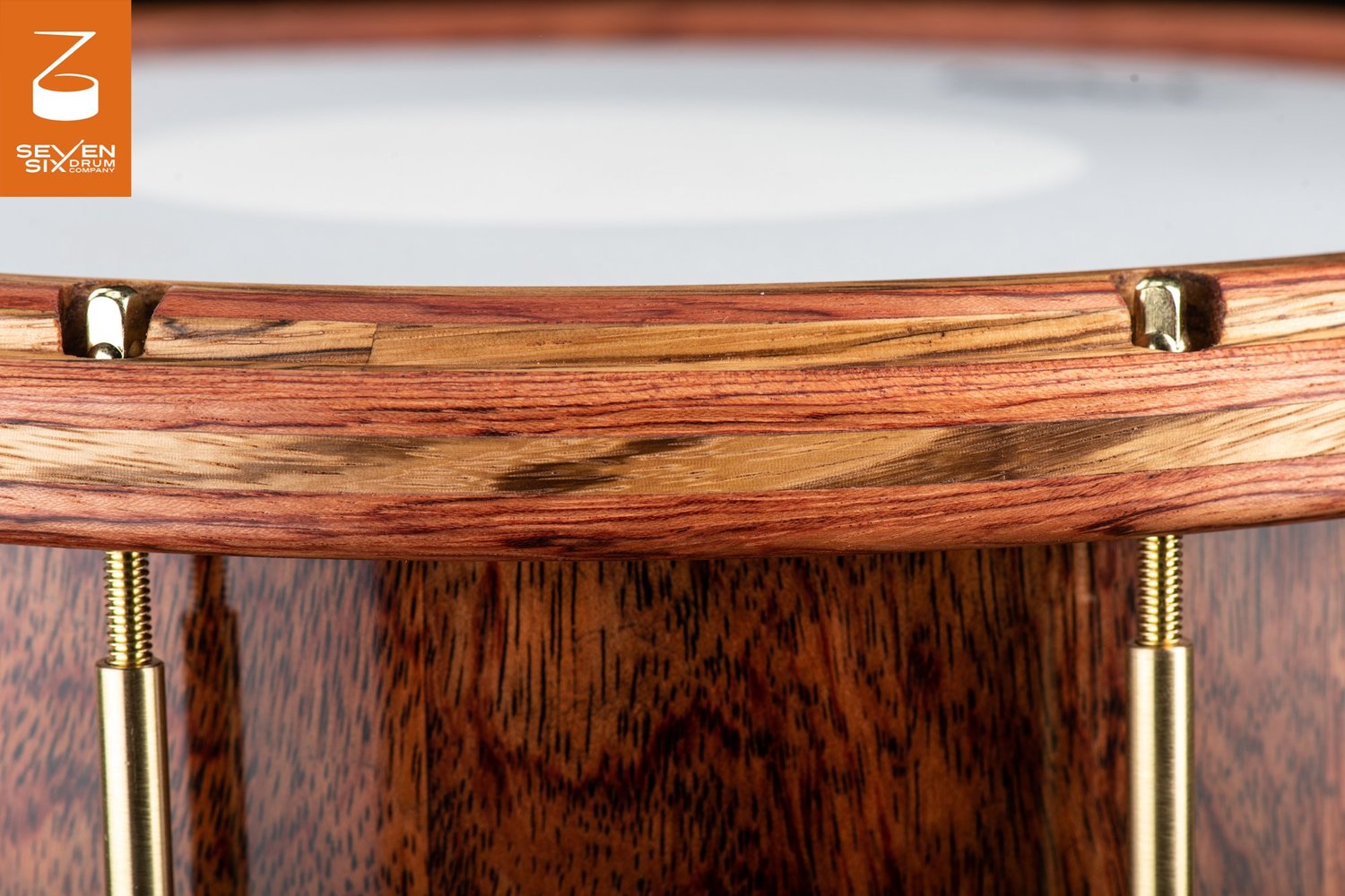 Close-up of a snare drum with a striped natural wood hoop, glossy grain wood shell, and brass tension rods.