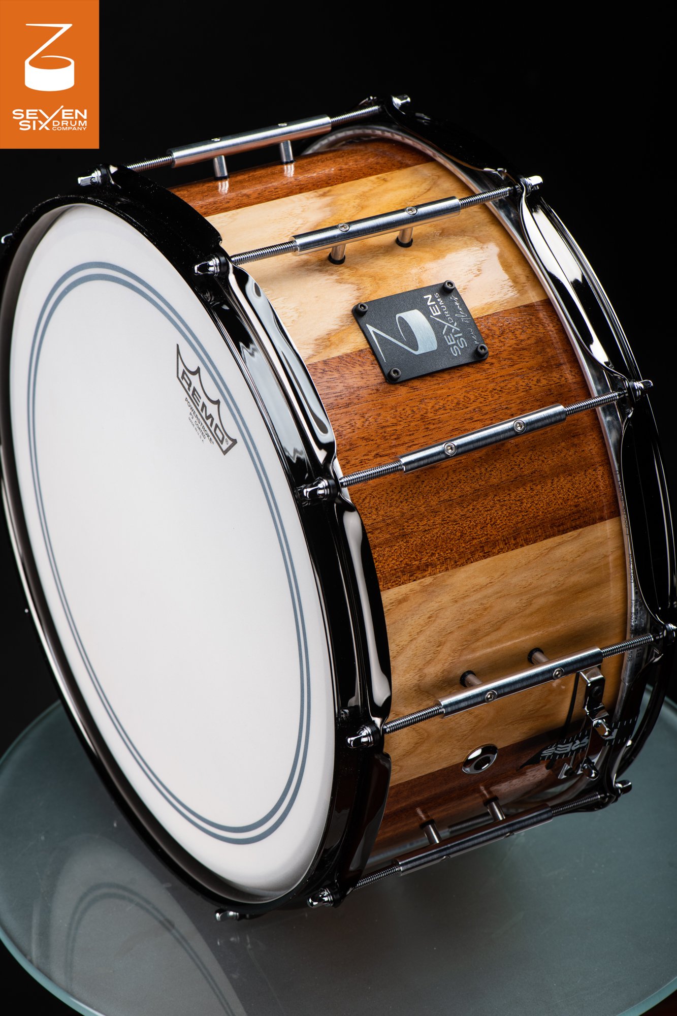 Handcrafted snare drum with a glossy striped light and dark wood shell, chrome hoops, and long metal tube lugs.