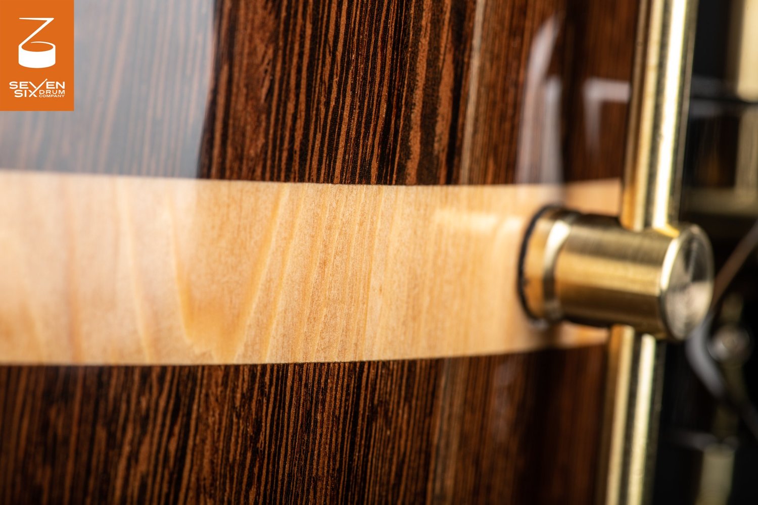 Detail of a high-gloss dark wood snare drum shell with a light wood center inlay and a brass tube lug.