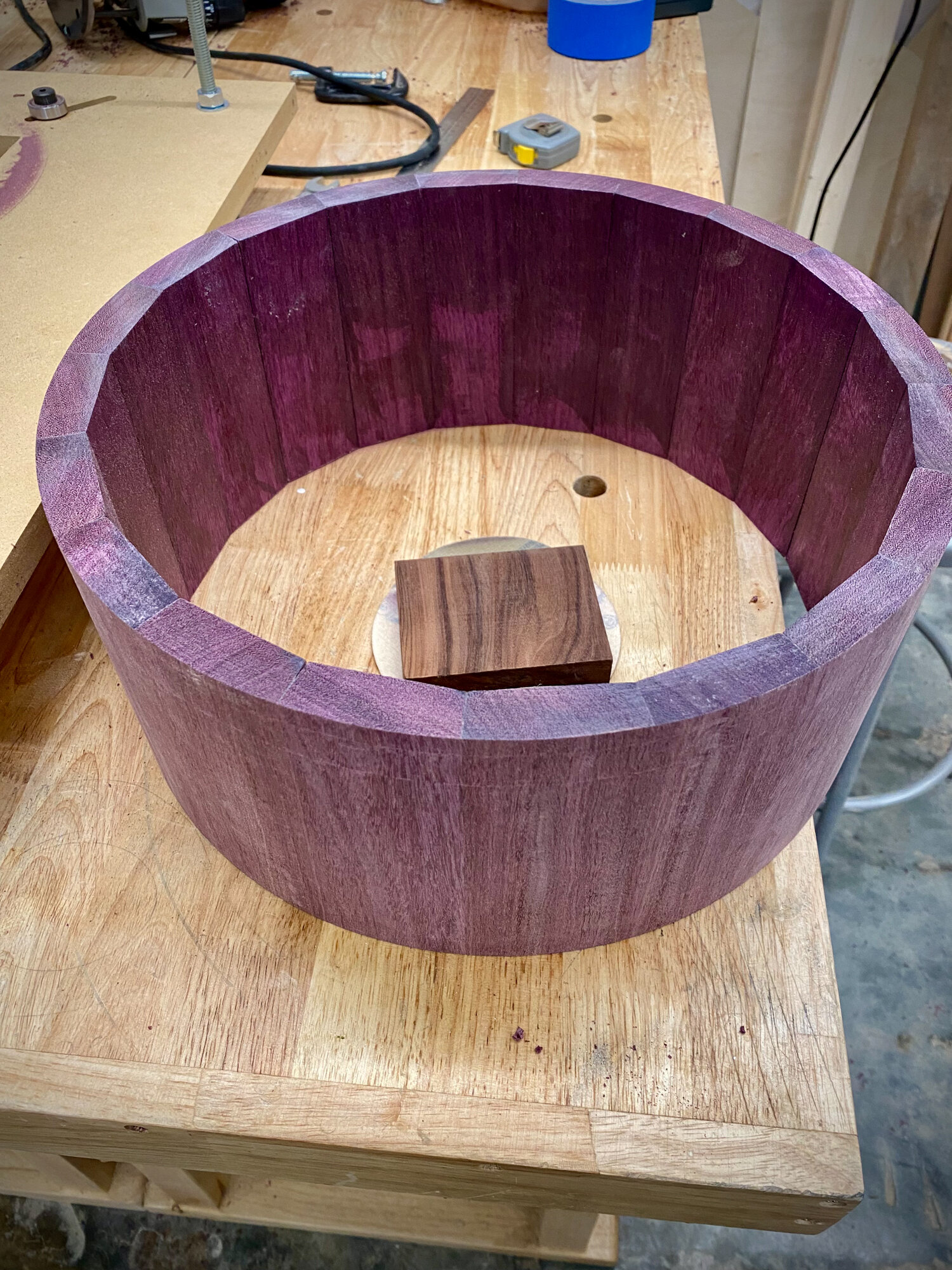 An unfinished purpleheart stave snare drum shell sits on a wooden workbench with no hardware installed.