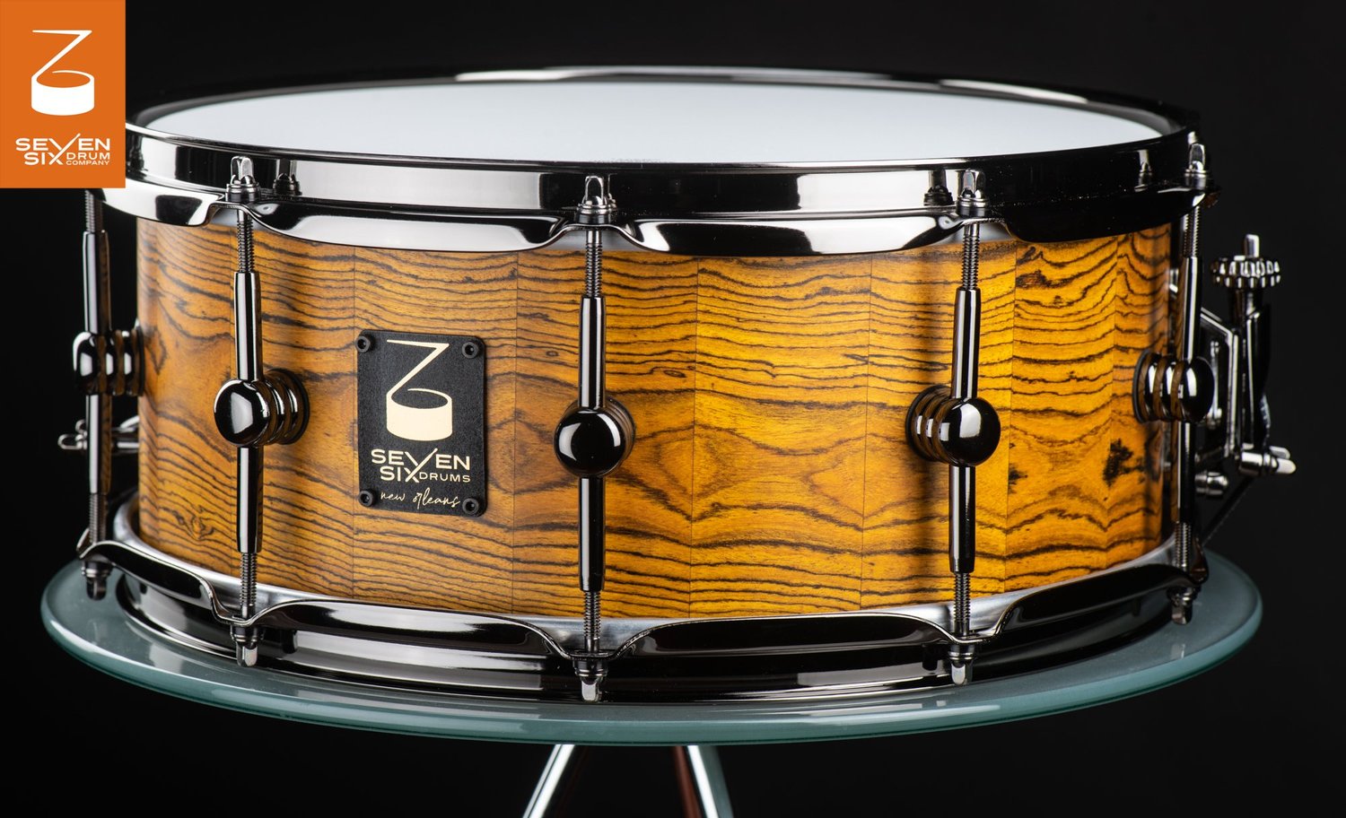 Handmade snare drum with an amber wood grain shell, black nickel hoops, and single-point tube lugs.