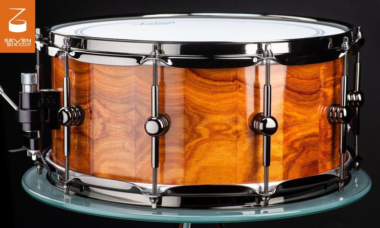 Handmade snare drum featuring a glossy amber wood shell with polished black nickel hoops and center-mounted lugs.