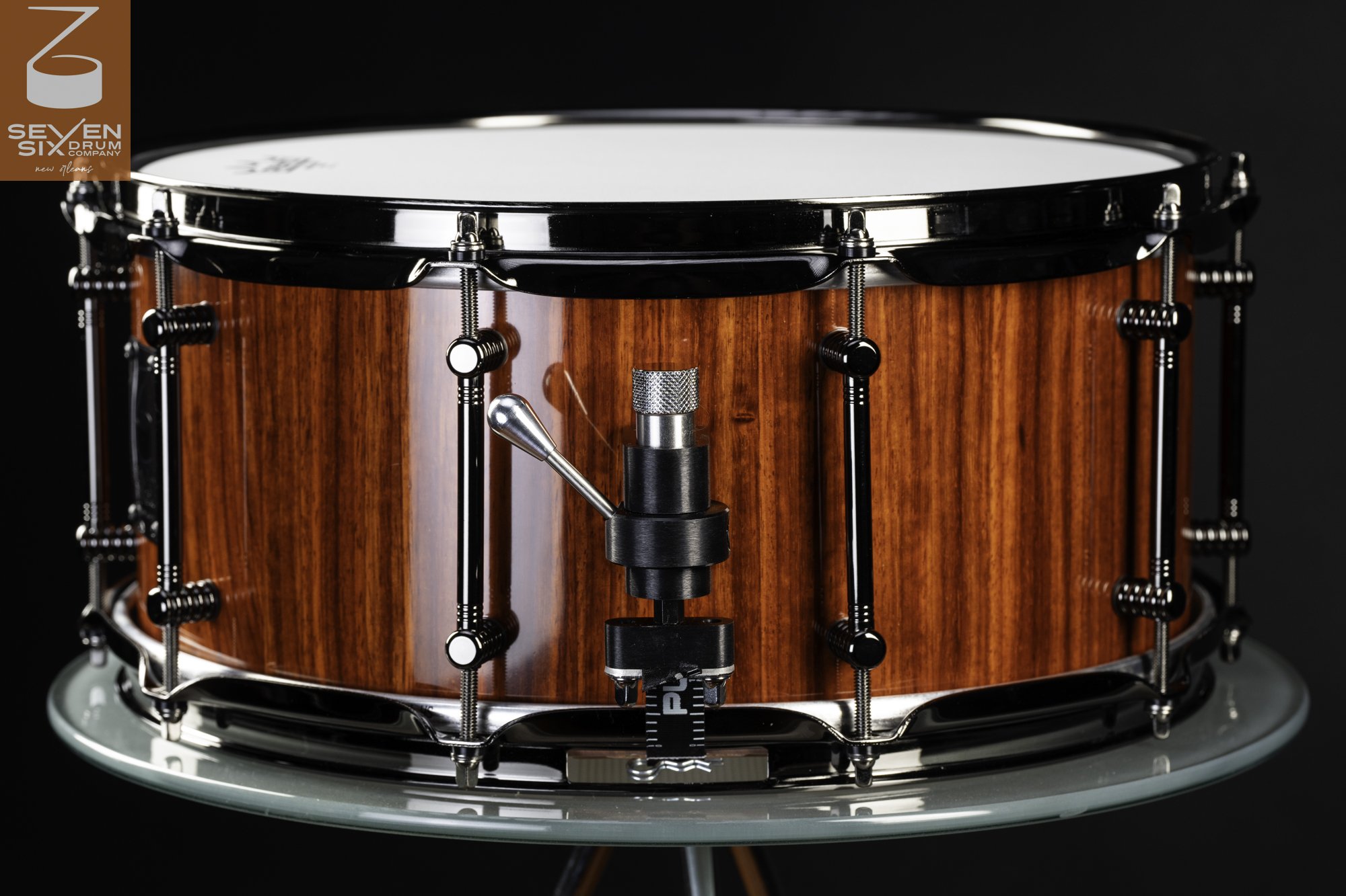 Handmade snare drum with a glossy reddish-brown wood shell, black nickel hoops, tube lugs, and a piston throw-off.