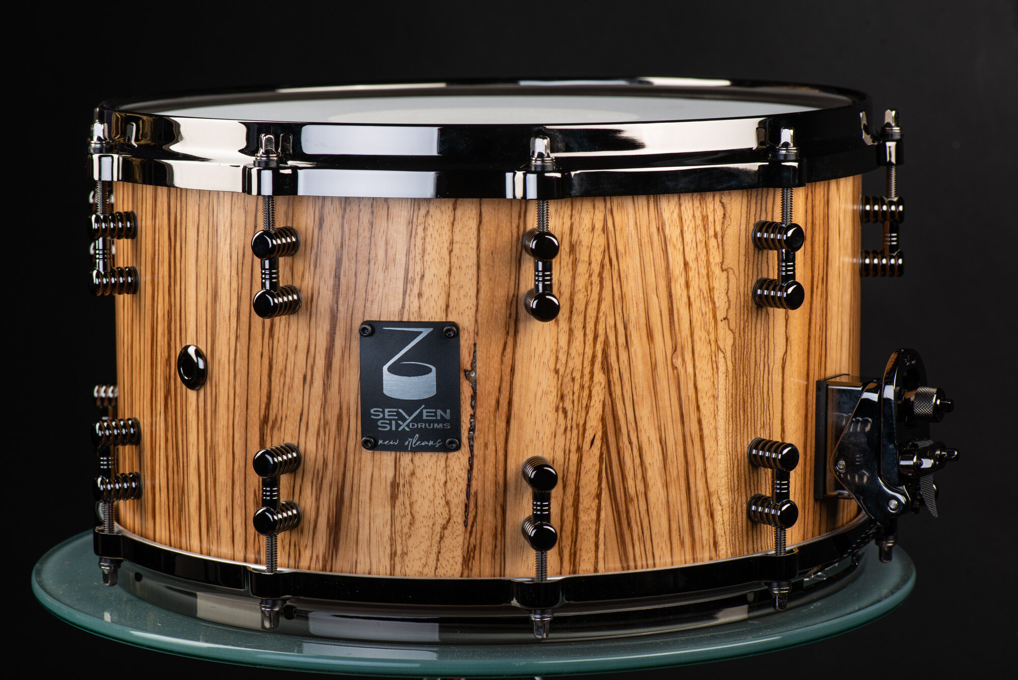 Handmade snare drum with a natural grain wood shell in a satin finish, fitted with black chrome hoops and lugs.