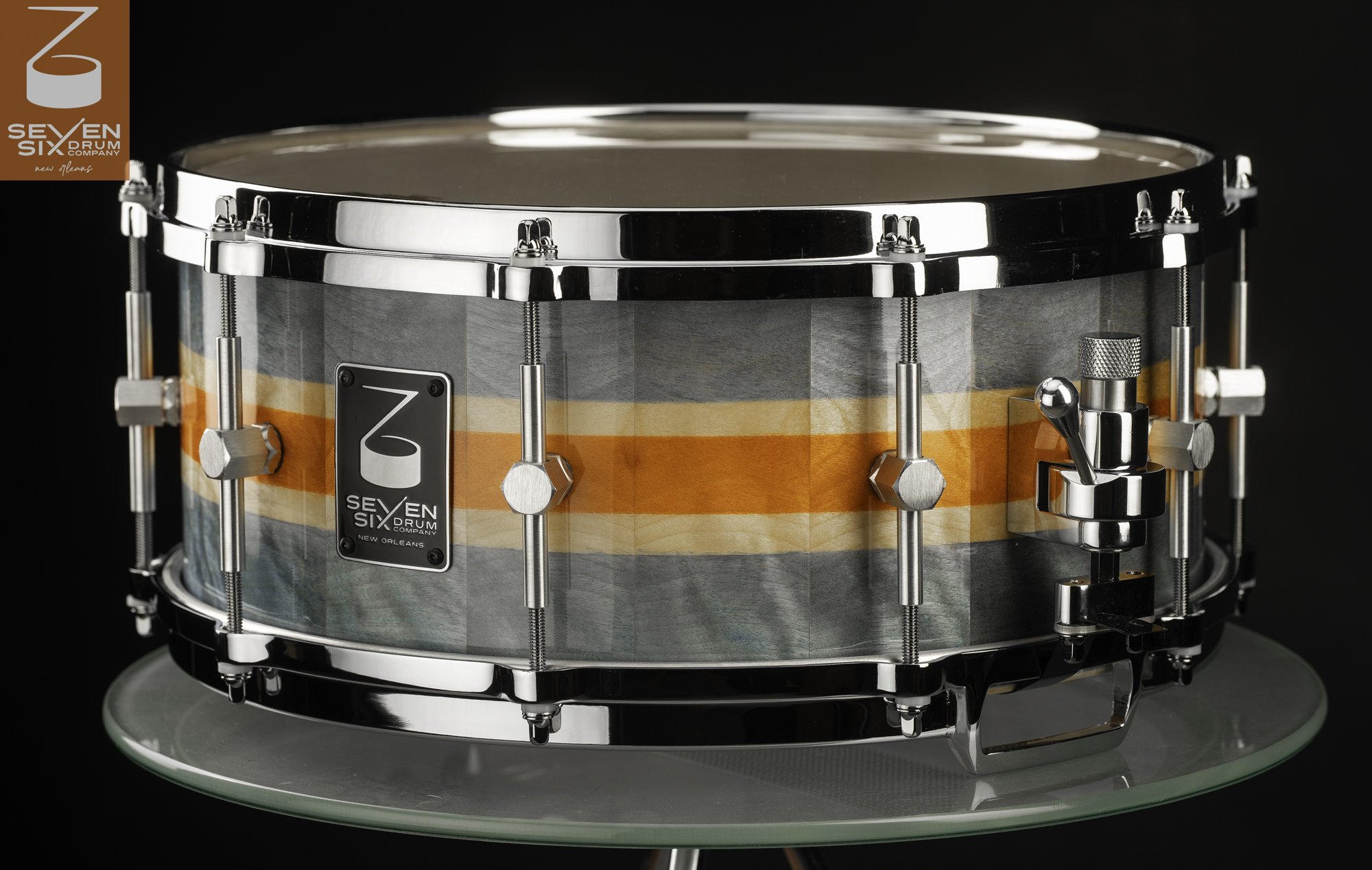 Handmade wood snare drum with glossy grey and orange stripes, fitted with chrome hoops, hex lugs, and a throw-off mechanism.