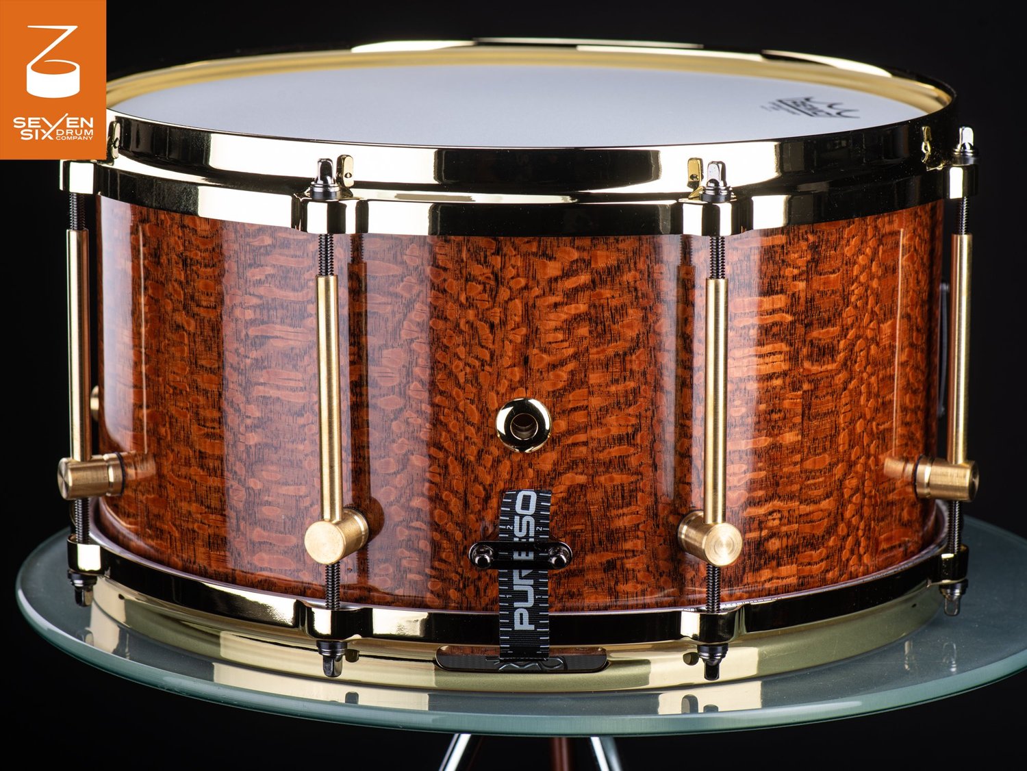 Handmade snare drum featuring a high-gloss figured wood shell with polished brass hoops and tube lugs.