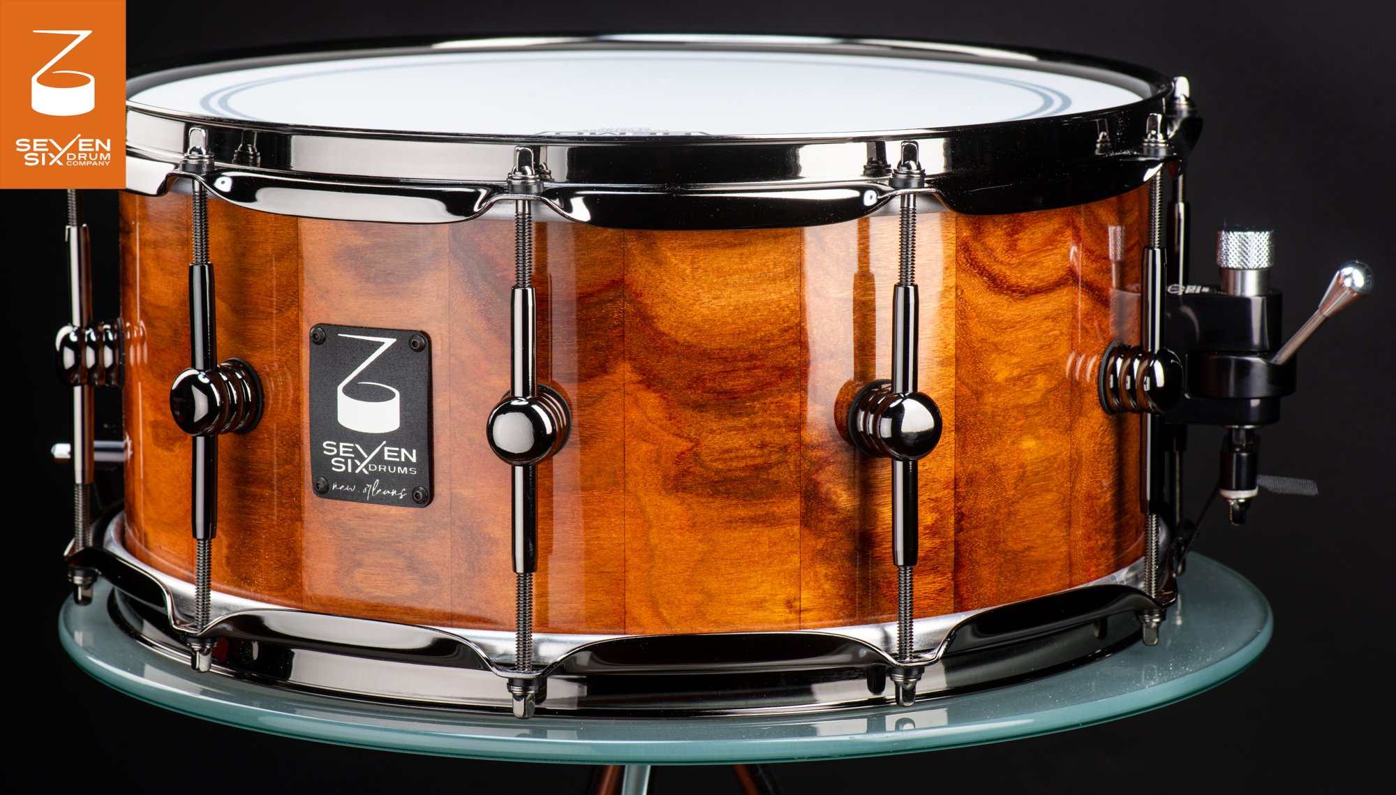 Handcrafted snare drum featuring a high-gloss amber wood shell, black nickel hoops, and spherical center lugs.
