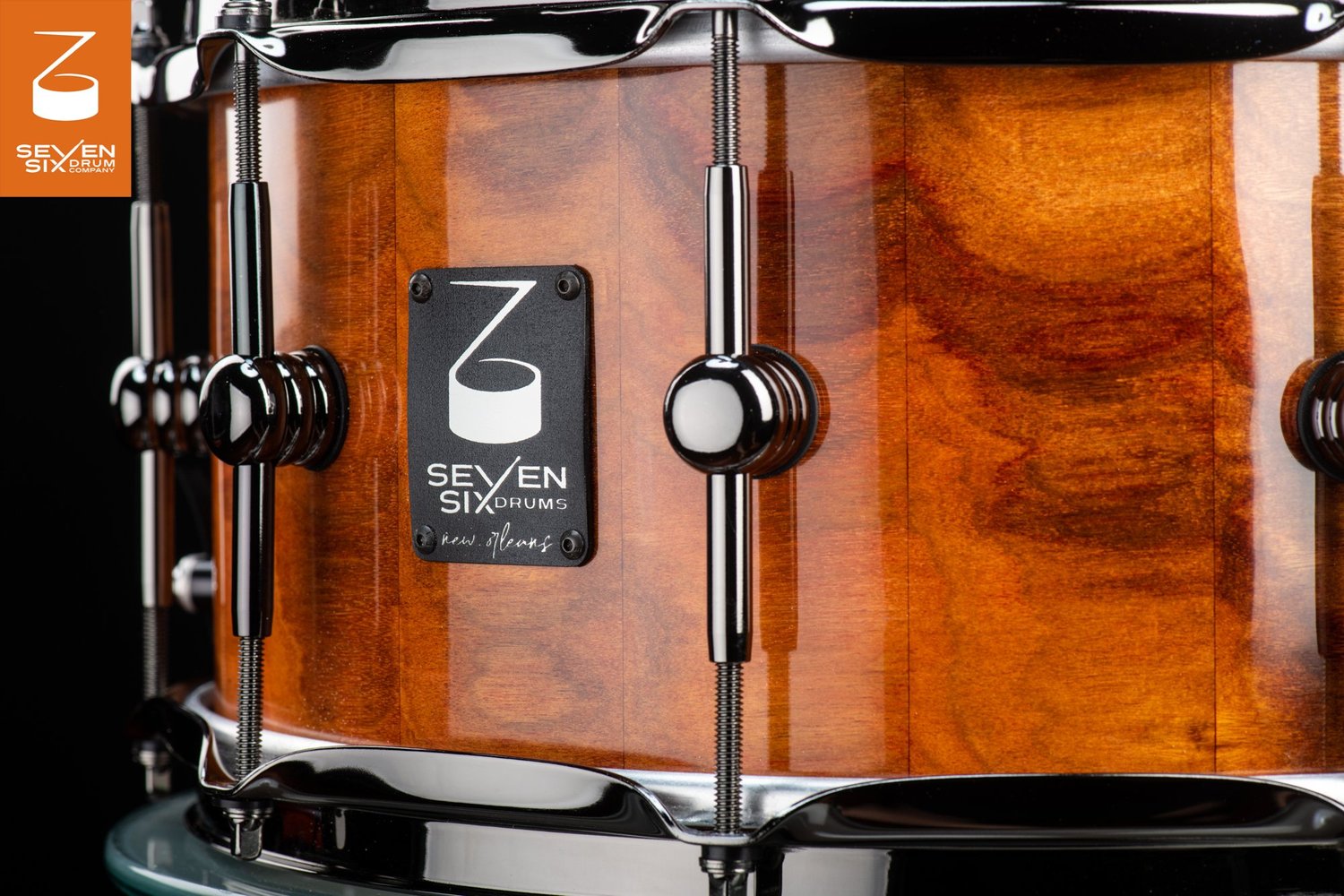 Snare drum with a glossy amber wood shell, black nickel lugs and hoops, and a black Seven Six Drums brand badge.