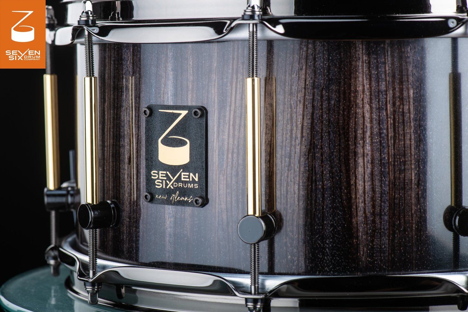 Snare drum with a dark vertical wood grain finish, black nickel hoops, and brass tube lugs with black mounts.