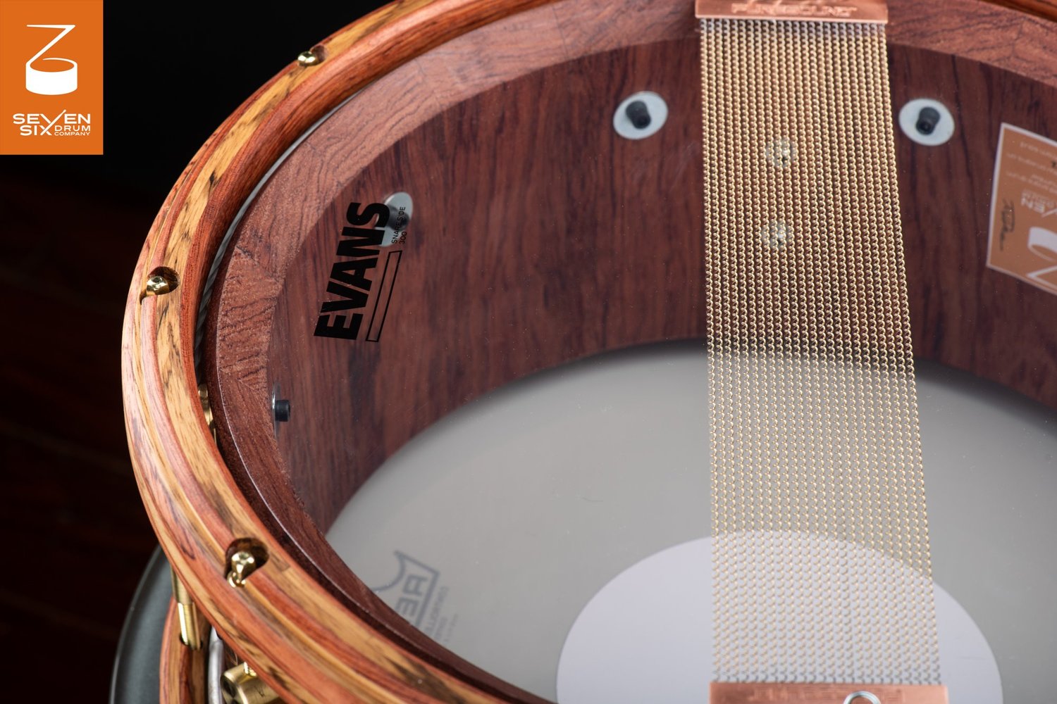 Handmade wood snare drum with a natural dark shell, striped wooden hoop, and brass-colored snare wires over a clear Evans head.