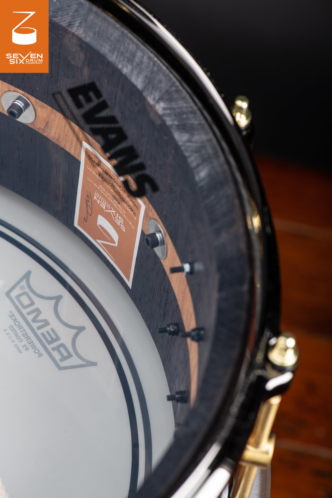 Interior of a snare drum with a dark textured wood shell, natural wood stripe inlay, black hoop, and polished brass lugs.