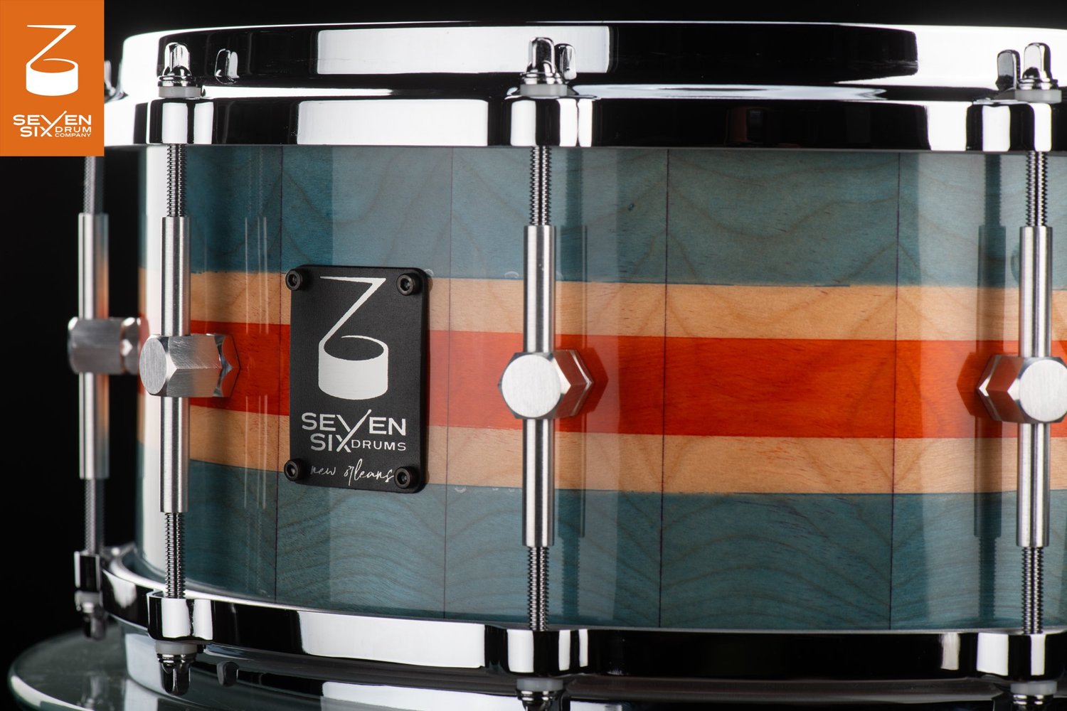 Wooden snare drum with glossy blue, natural, and orange stripes, polished chrome hoops, and hexagonal lugs.