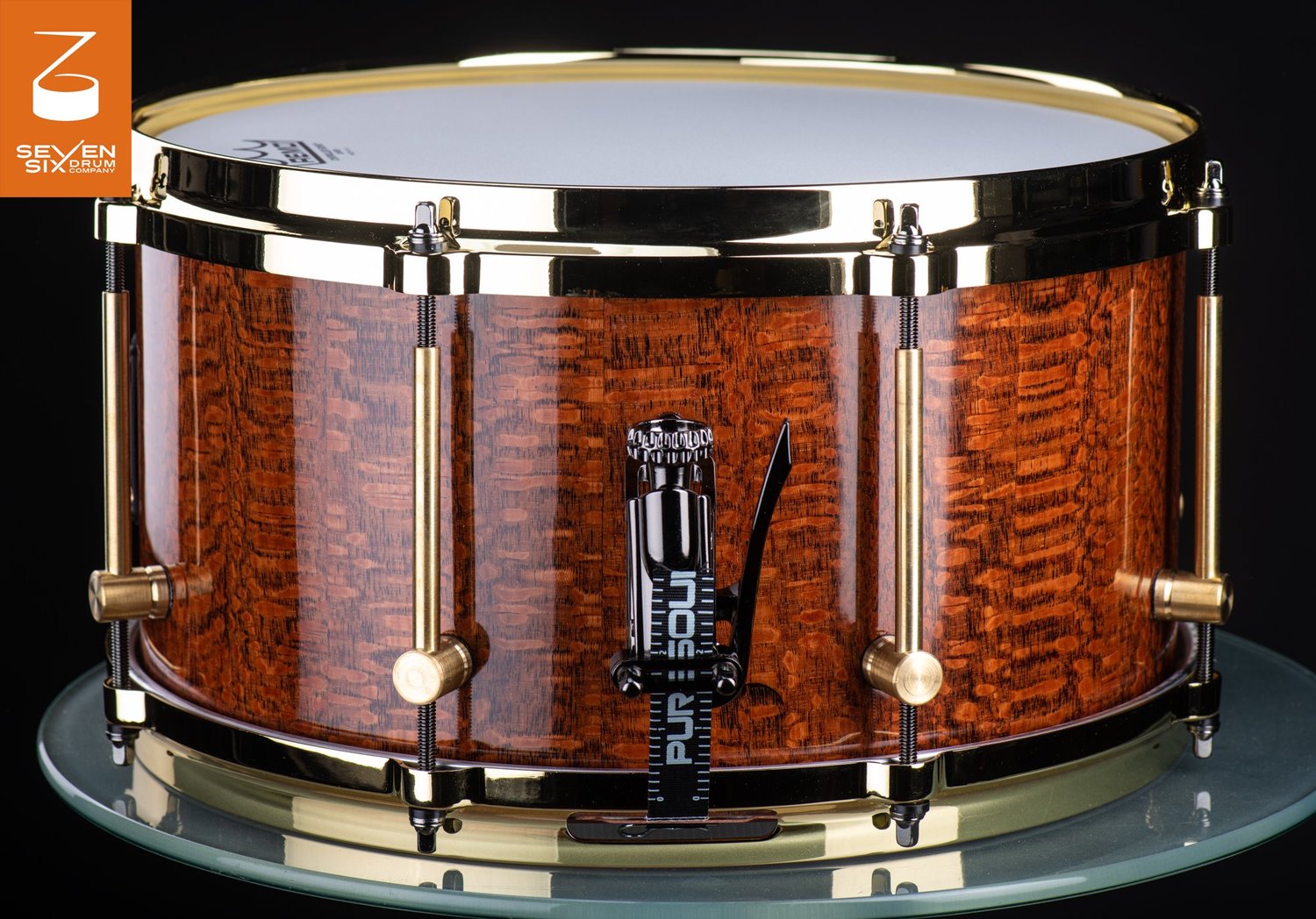 Glossy amber figured-wood snare drum with polished brass hoops, brass tube lugs, and a black snare throw-off.