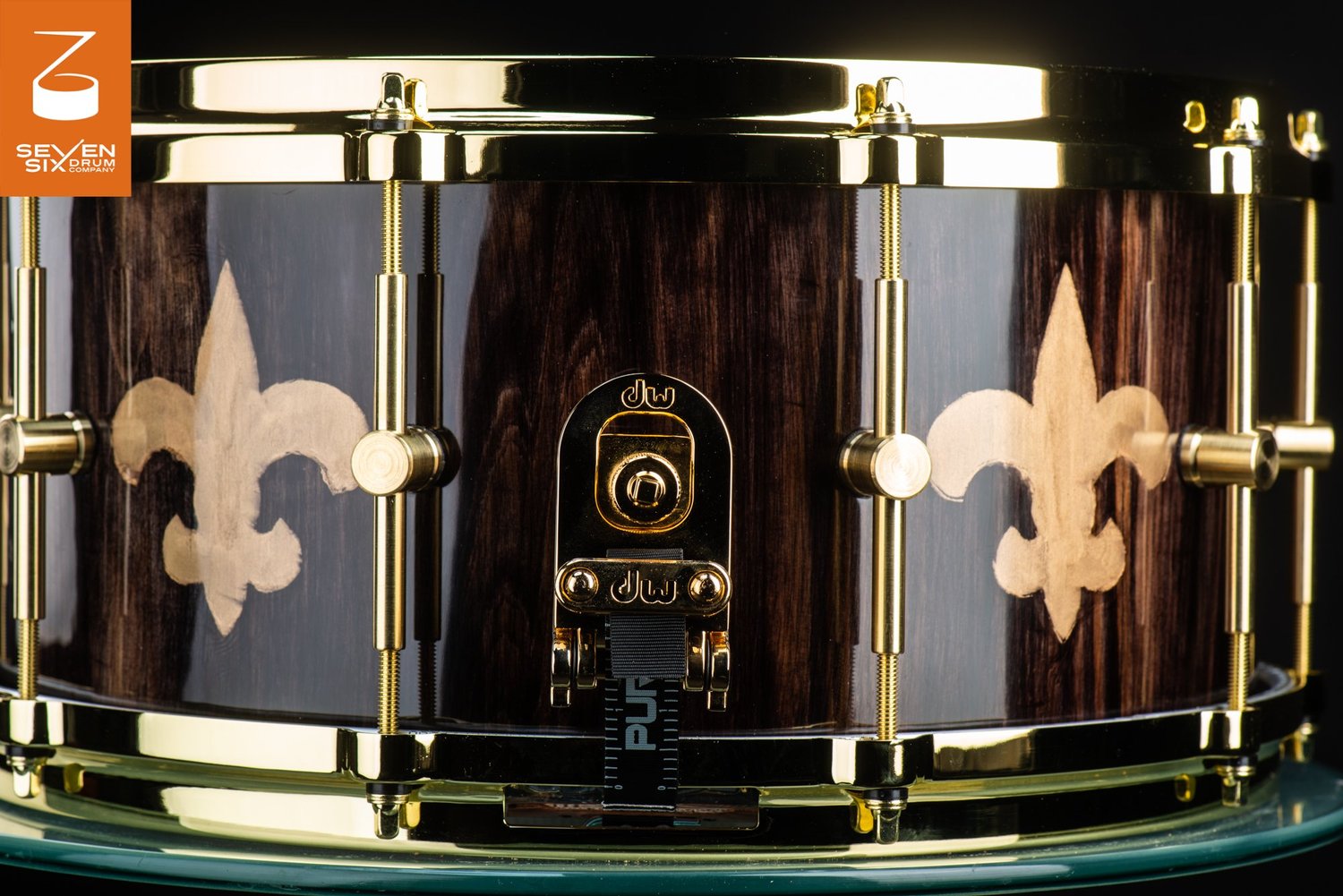A glossy dark wood snare drum featuring fleur-de-lis inlays, polished gold rims, tube lugs, and a gold butt plate.