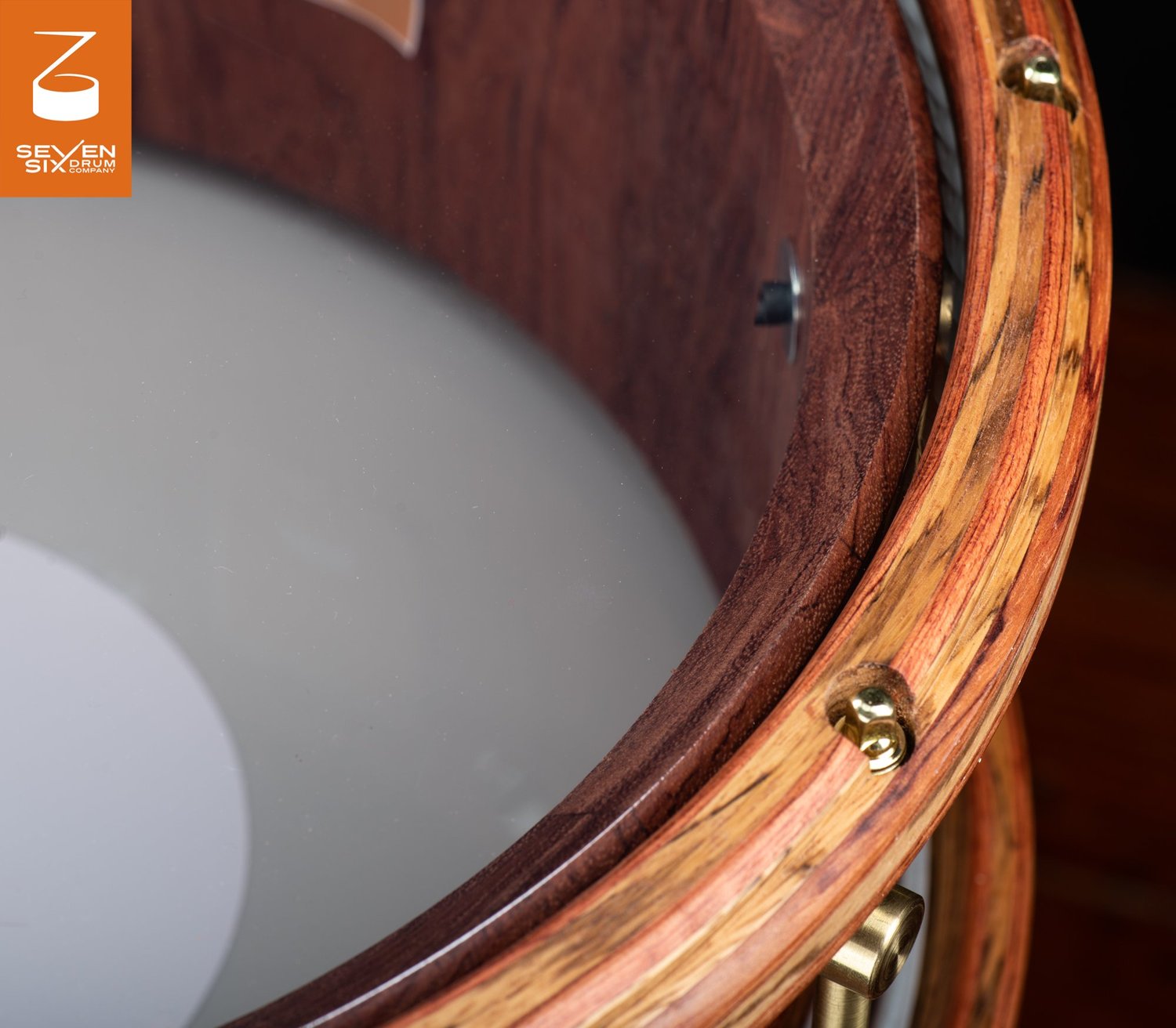 A handmade dark wood snare drum featuring a natural-finish striped wooden hoop secured by recessed brass hardware.