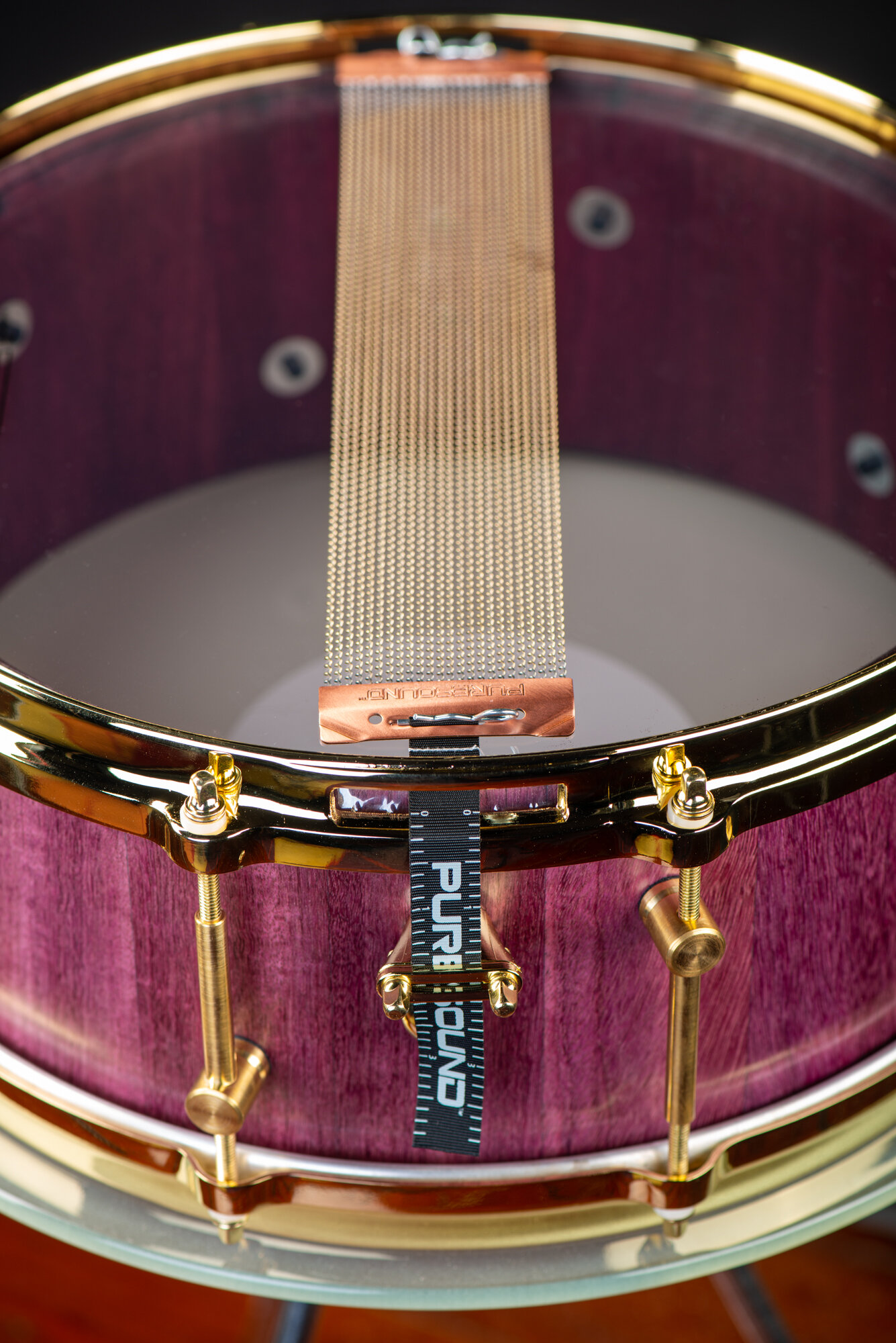 Purpleheart wood snare drum showing polished gold hoops, brass tube lugs, and PureSound snare wires over a clear resonant head.