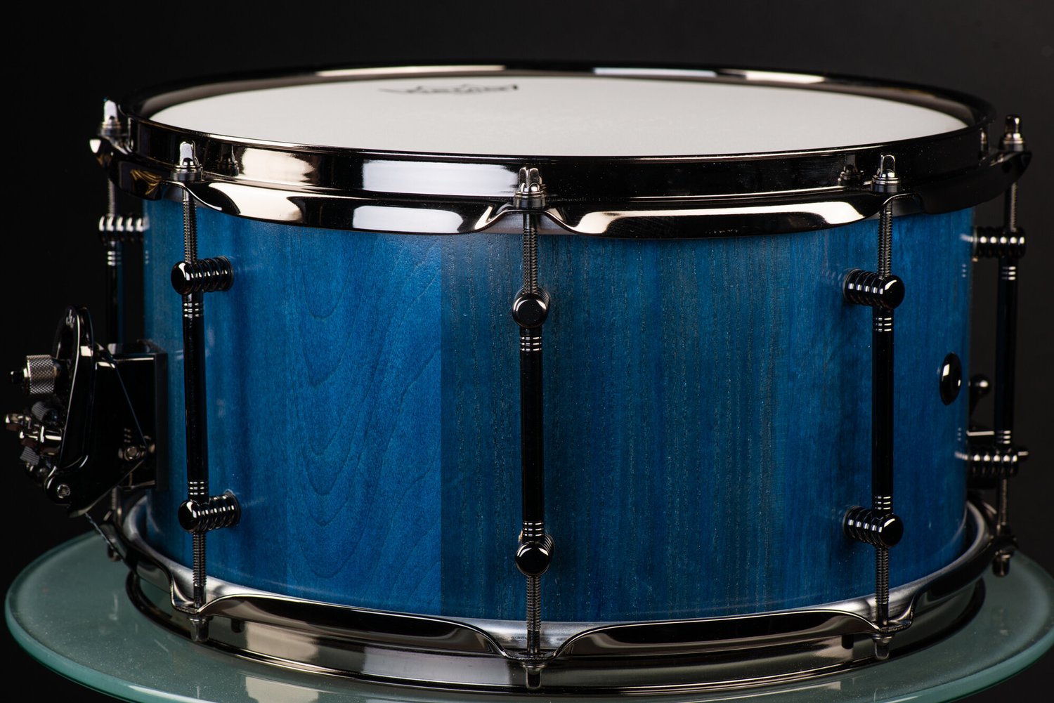 Handmade snare drum featuring a blue-stained wood shell, satin finish, and polished black nickel tube lugs and hoops.