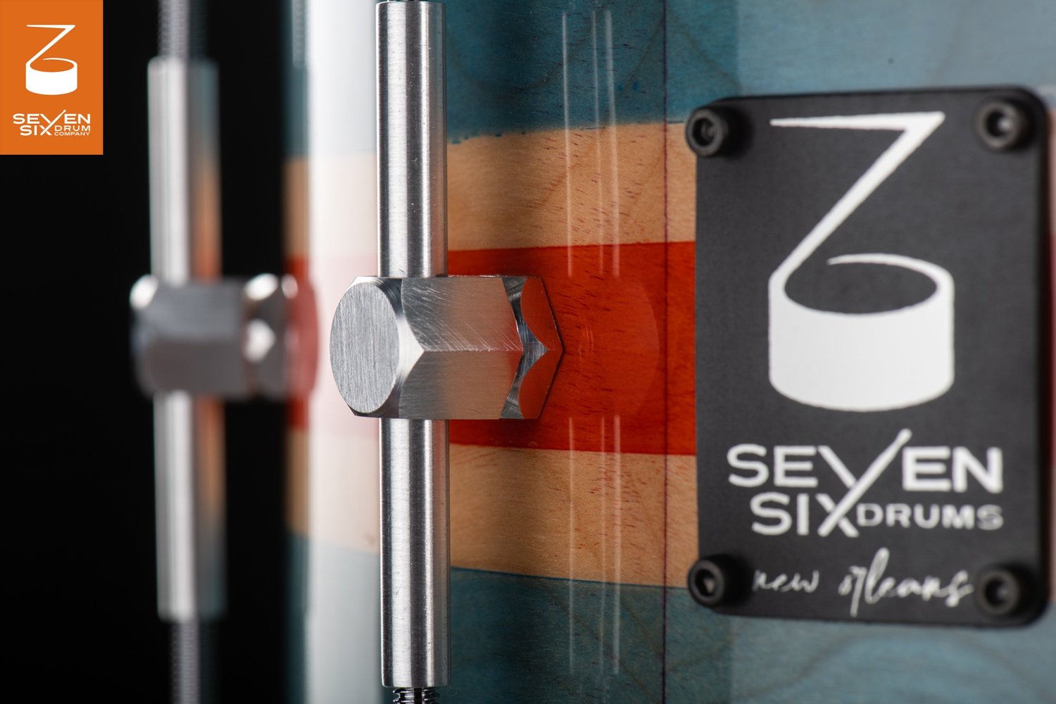 Close-up of a gloss-finished striped wood snare drum featuring a brushed metal tube lug and black brand badge.