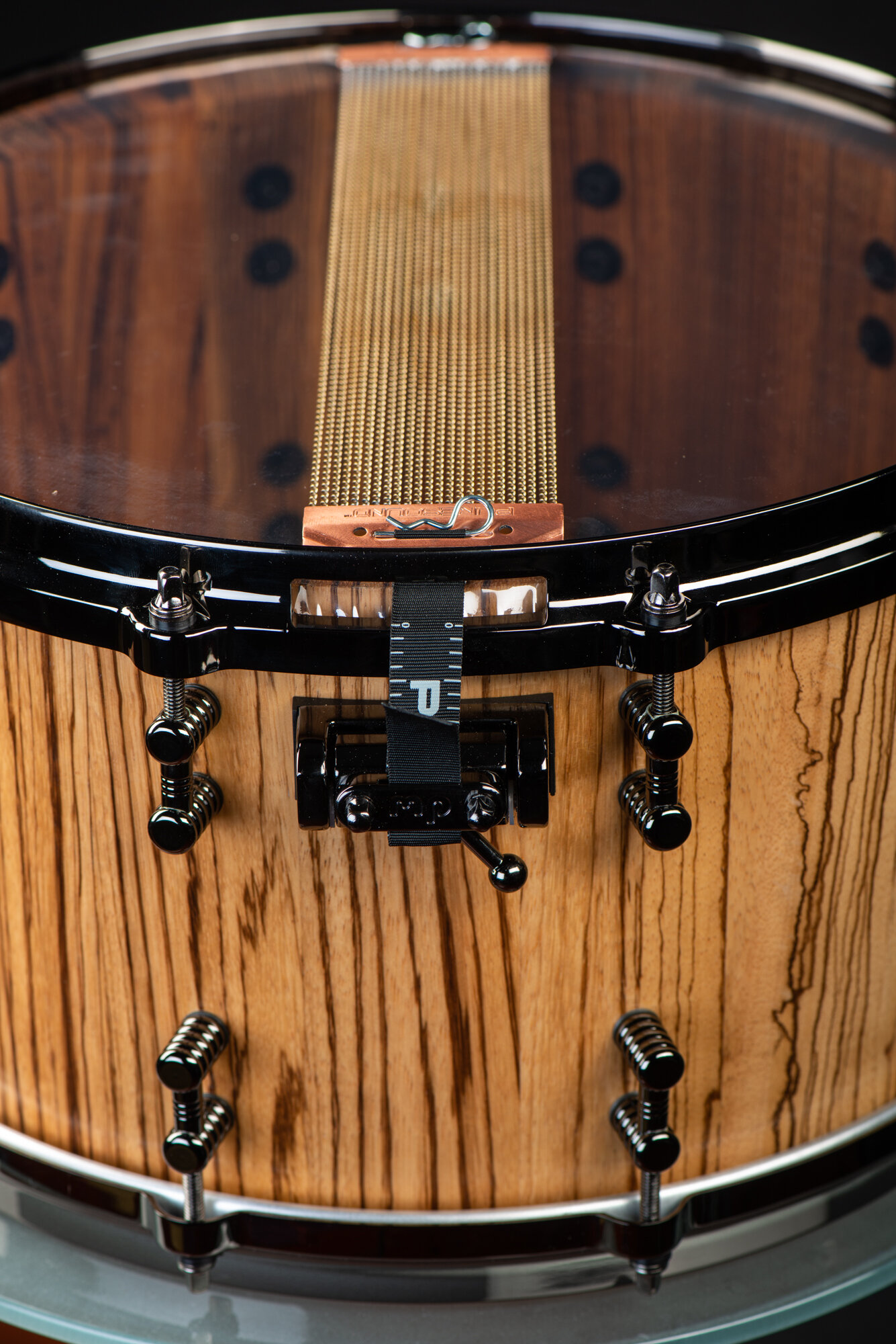 Natural finish zebrawood snare drum featuring gloss black hardware, dual lugs, and brass snare wires on a clear resonant head.