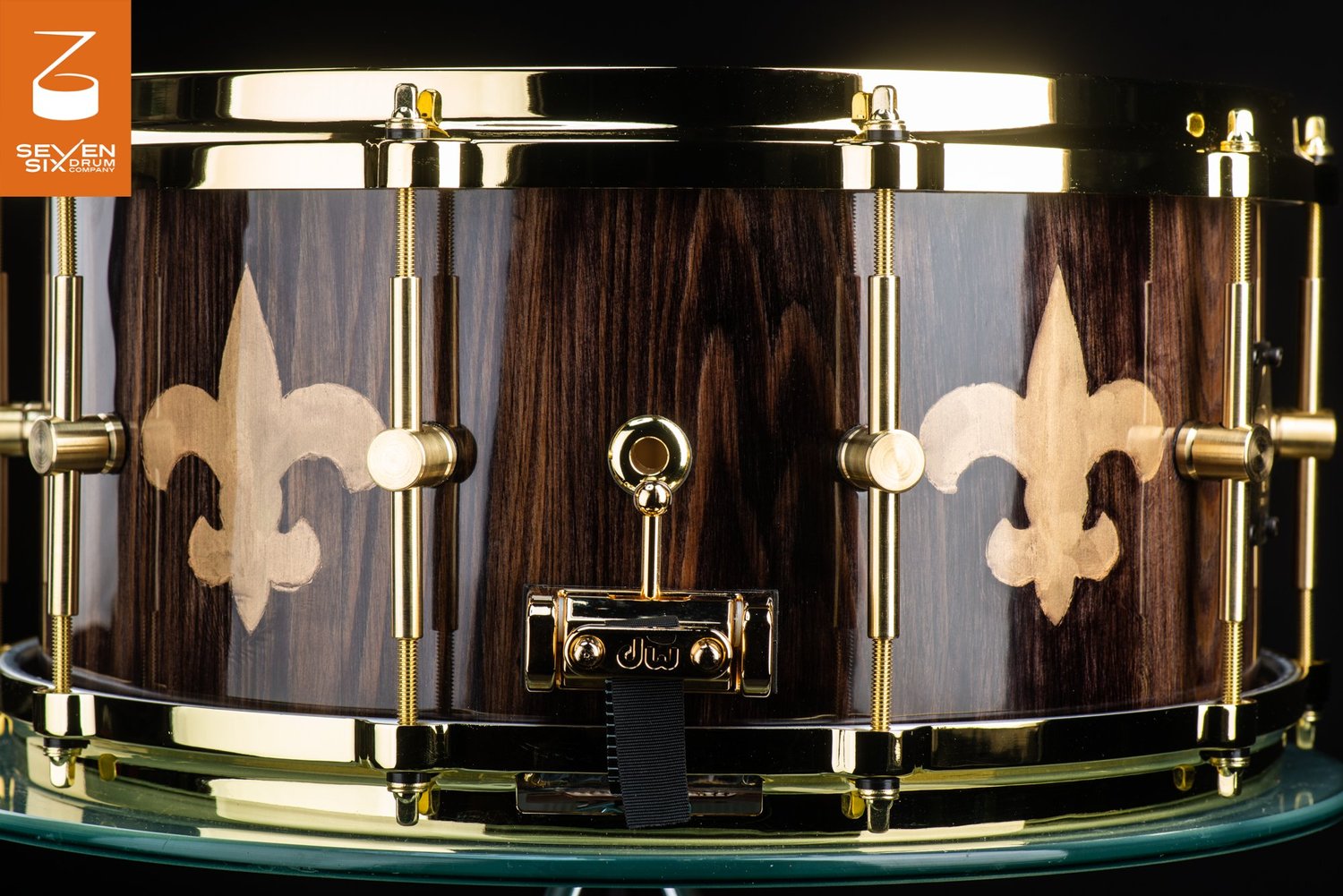 Snare drum with a high-gloss dark wood grain shell, inlaid fleur-de-lis, and polished brass tube lugs and hoops.