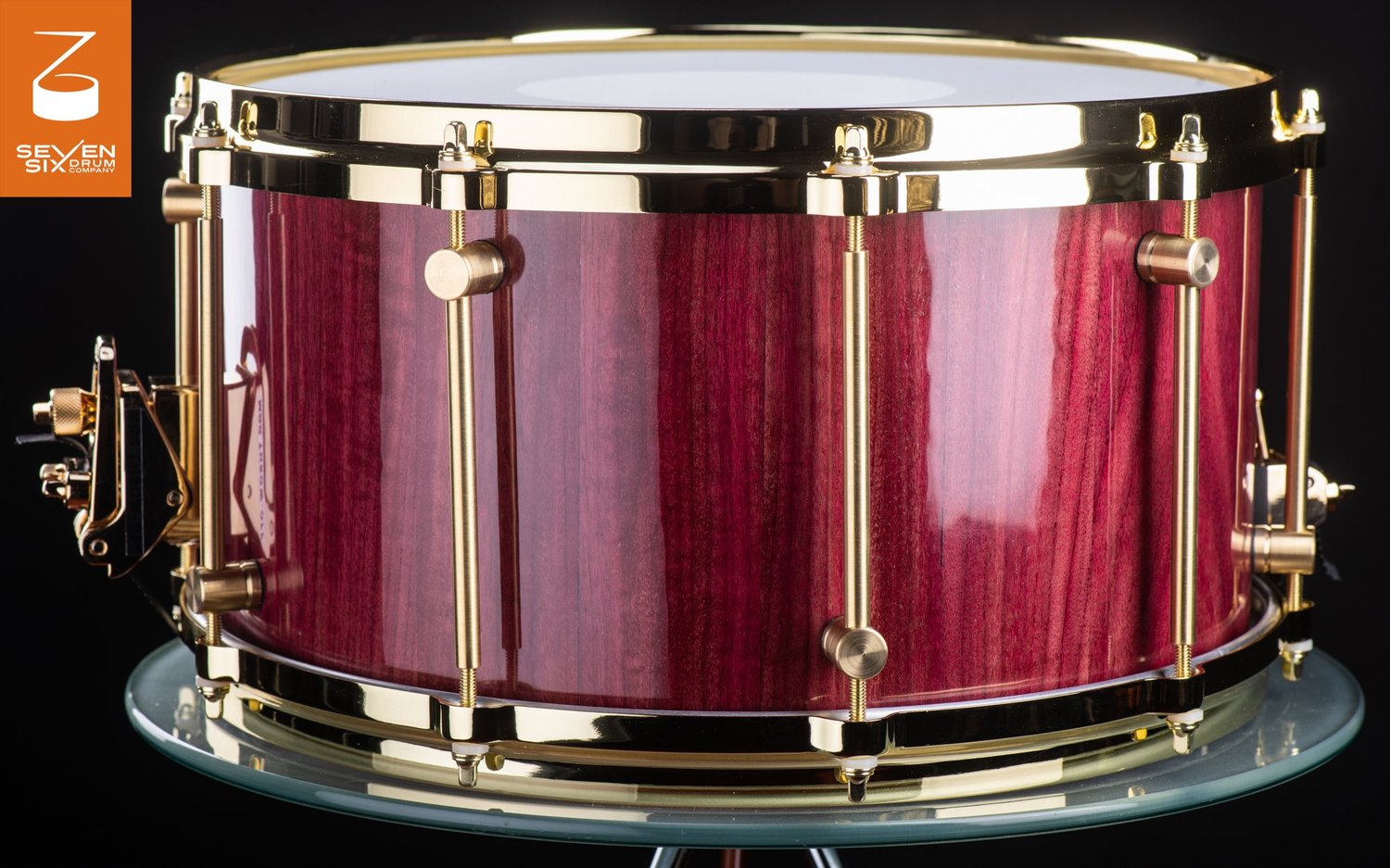 Handcrafted snare drum featuring a glossy purpleheart shell, polished brass tube lugs, and matching gold hoops.