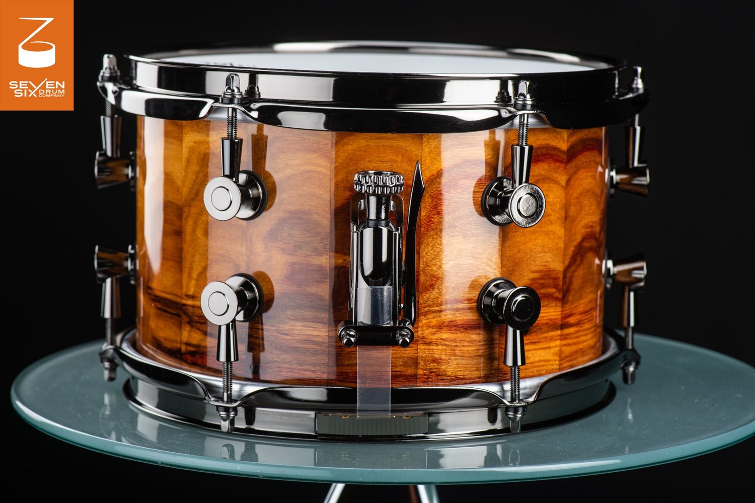 Handmade snare drum with a glossy amber wood stave shell, black nickel hoops, round lugs, and a snare throw-off.