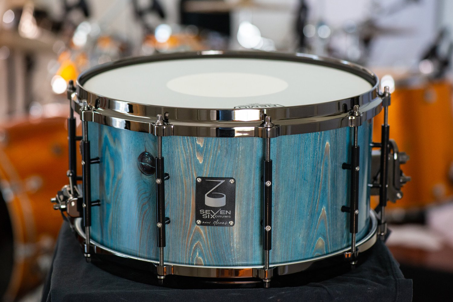Wooden snare drum featuring a blue-stained wood grain shell, black nickel hoops, and tube lugs.