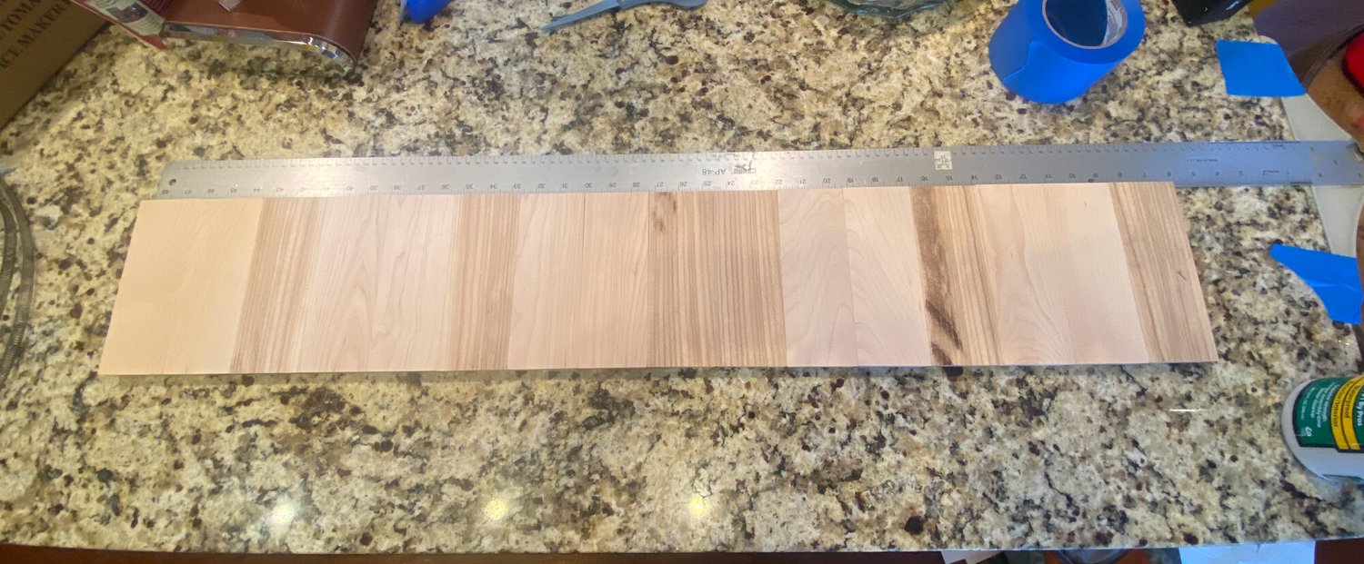 Unfinished light wood staves laid flat next to a long metal ruler to measure for a snare drum shell.