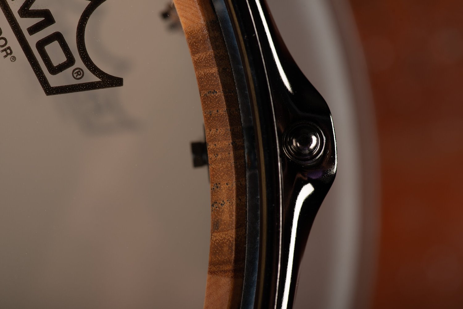 Close-up of a snare drum with a natural segmented wood shell, Remo head, and polished black chrome hoop with a tension rod.