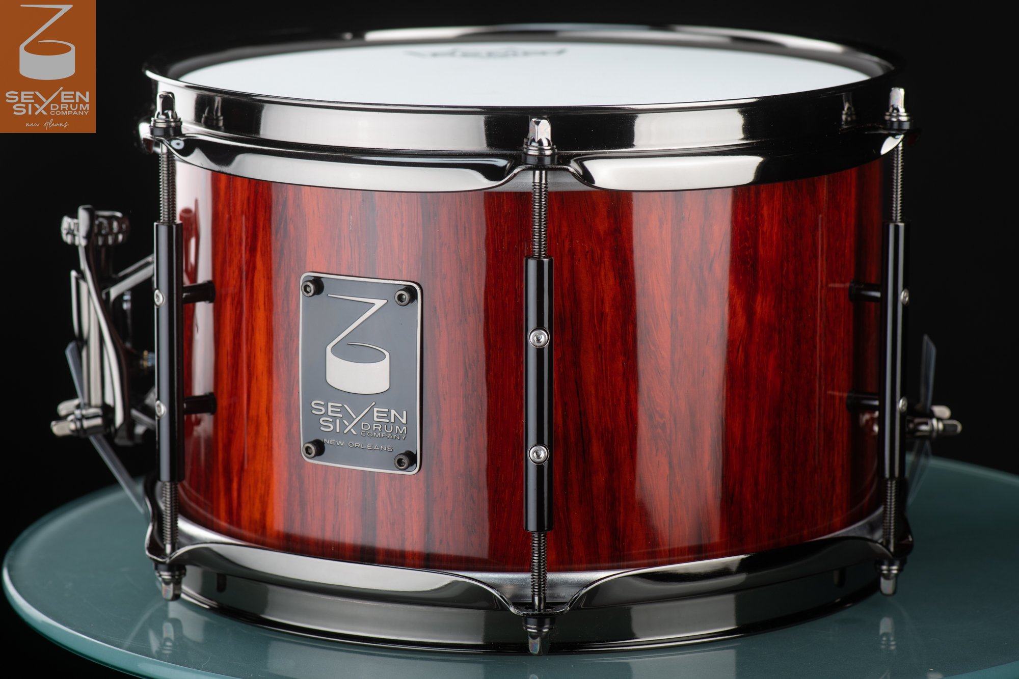 Handmade snare drum with a glossy reddish-brown wood shell, black nickel hoops, and tube lugs.