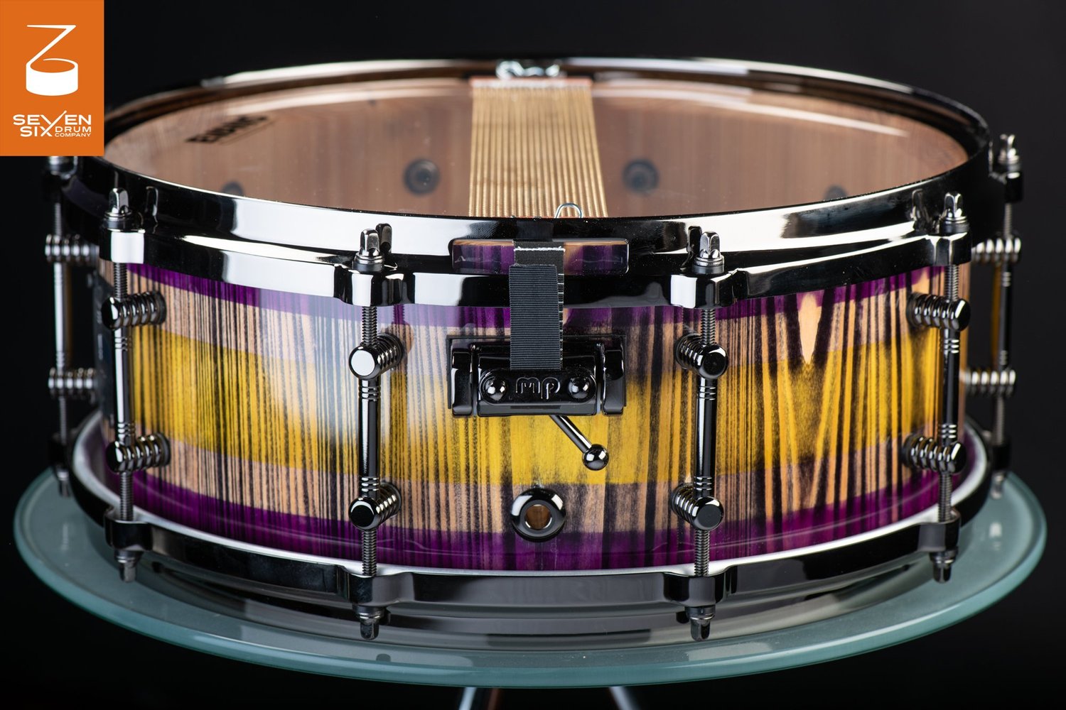 Snare drum featuring a purple and yellow striped wood shell with a gloss finish, black nickel hoops, tube lugs, and throw-off.