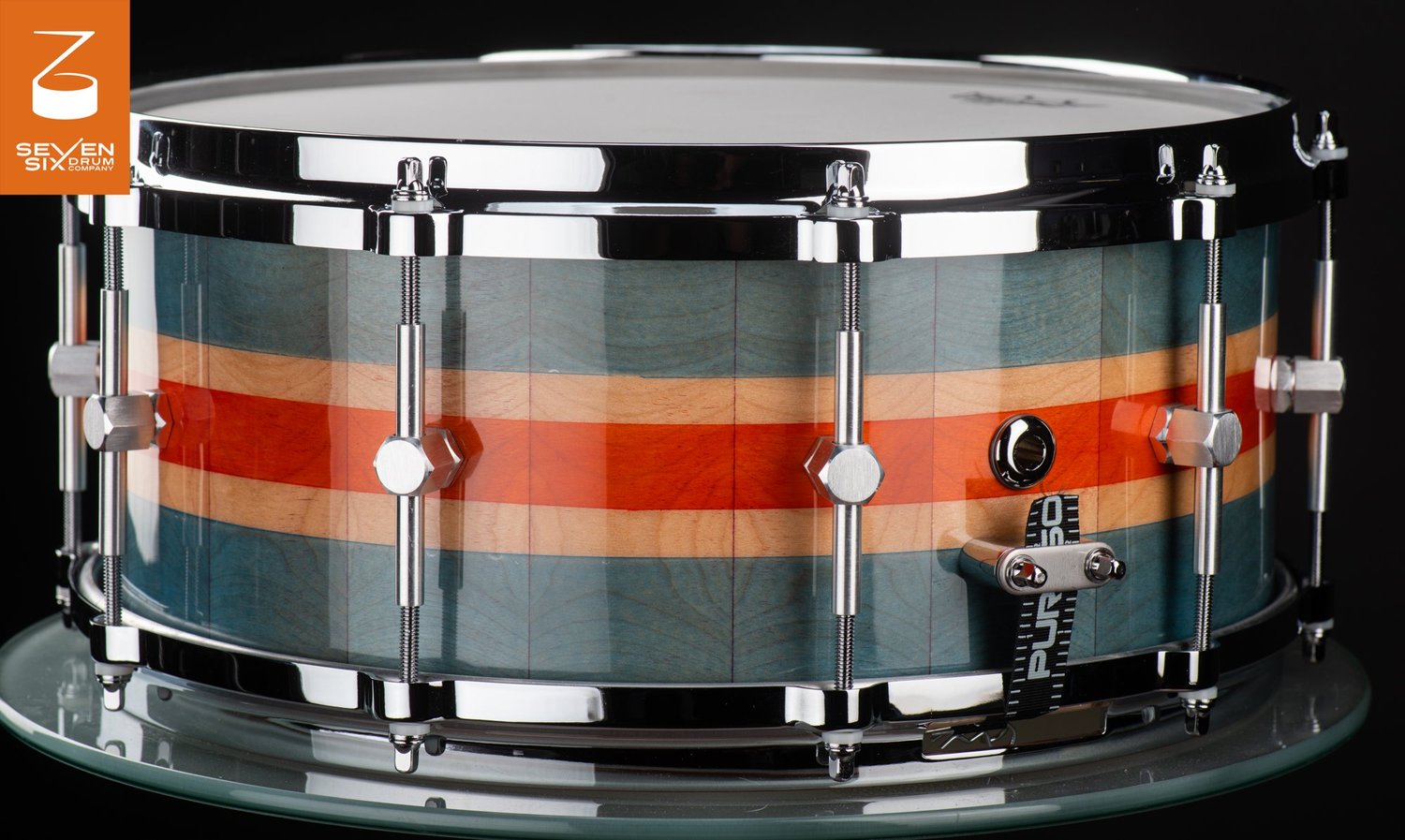 Glossy striped wood snare drum featuring teal and orange bands, chrome hoops, and single-point hex lugs.