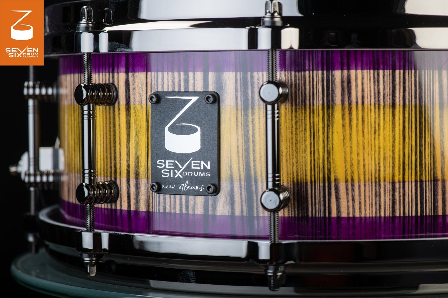 Snare drum featuring a glossy purple and yellow striped wood shell, black nickel hoops, and tube lugs.