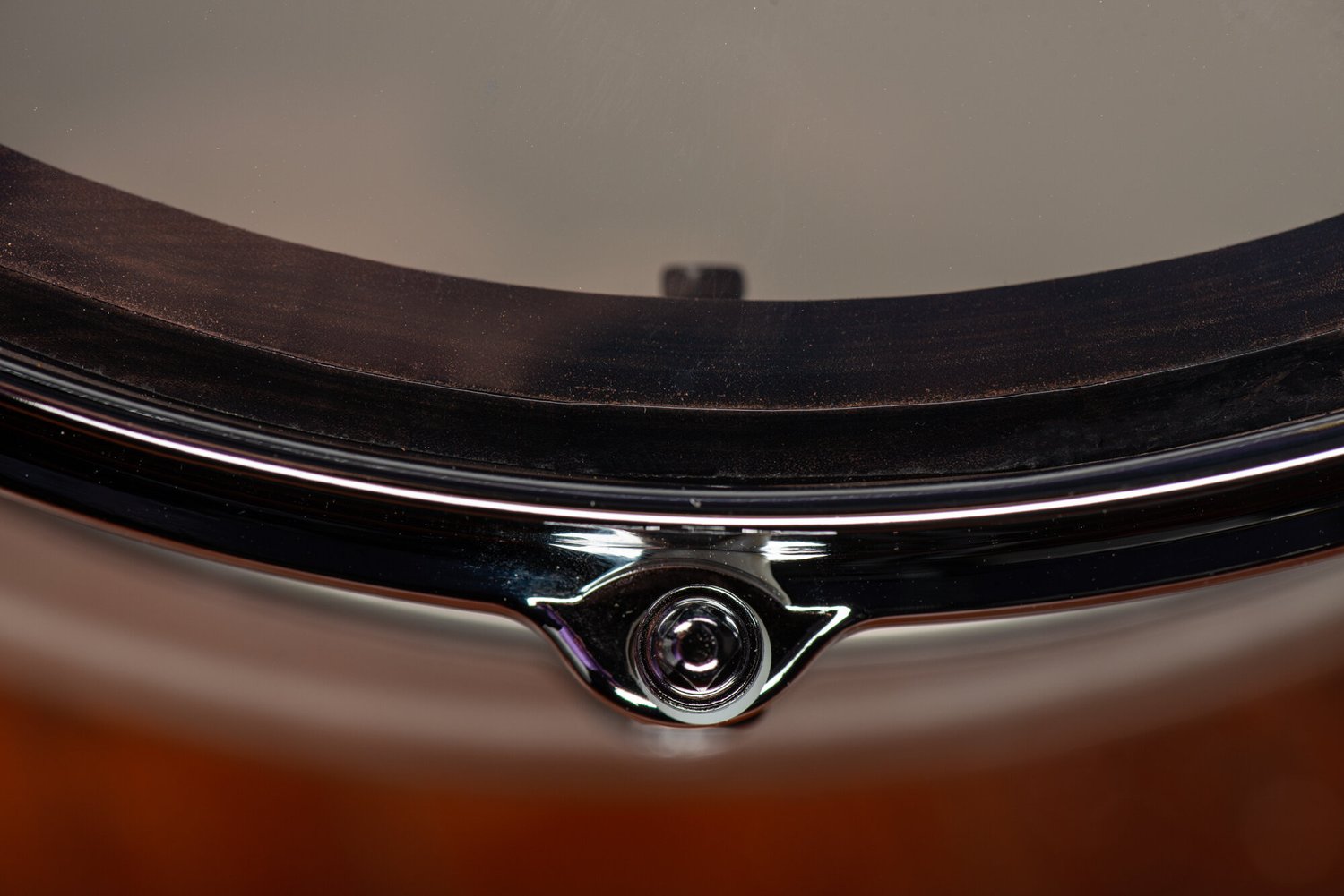 Snare drum detail featuring a polished chrome hoop, tension rod ear, and dark hardwood shell beneath a clear drumhead.