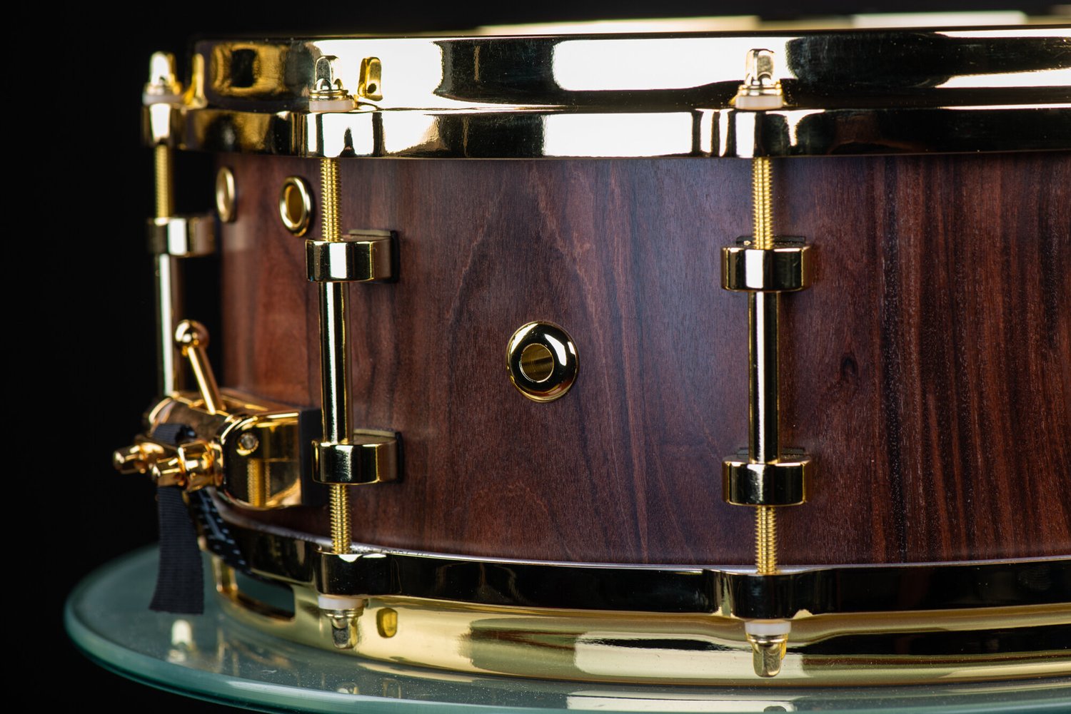 Handmade dark wood snare drum with a natural satin finish, fitted with polished gold hoops, lugs, and throw-off.