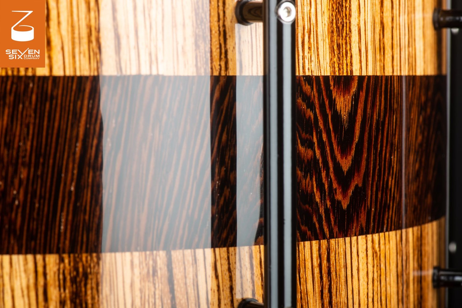 Close-up of a glossy zebrawood snare drum shell with a dark center band and a black tube lug.