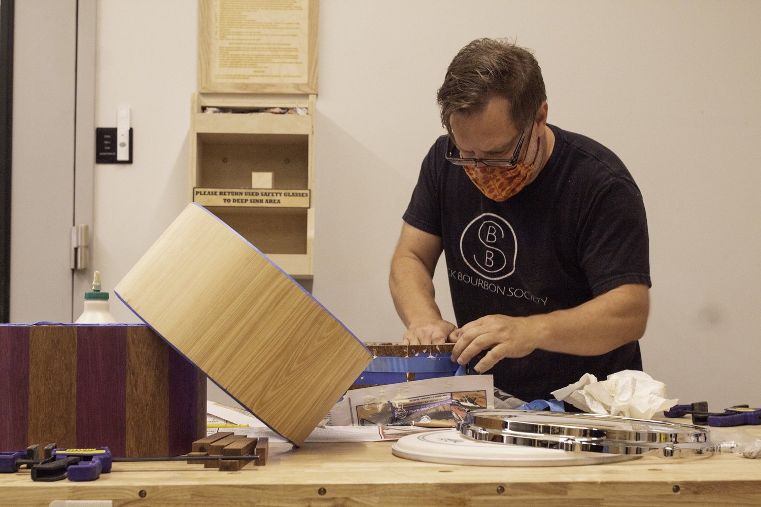A craftsman glues an unfinished wooden shell on a workbench beside striped stave shells and a polished chrome hoop.