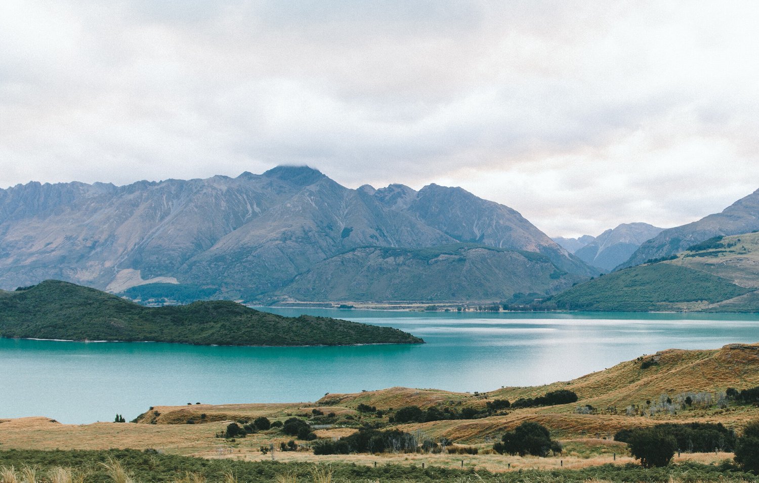 Turquoise lake surrounded by rolling grassy hills and rugged mountain peaks under an overcast sky.