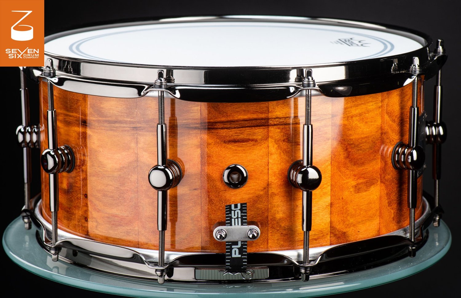 Handmade snare drum featuring a high-gloss amber wood stave shell fitted with black nickel hoops and tube lugs.