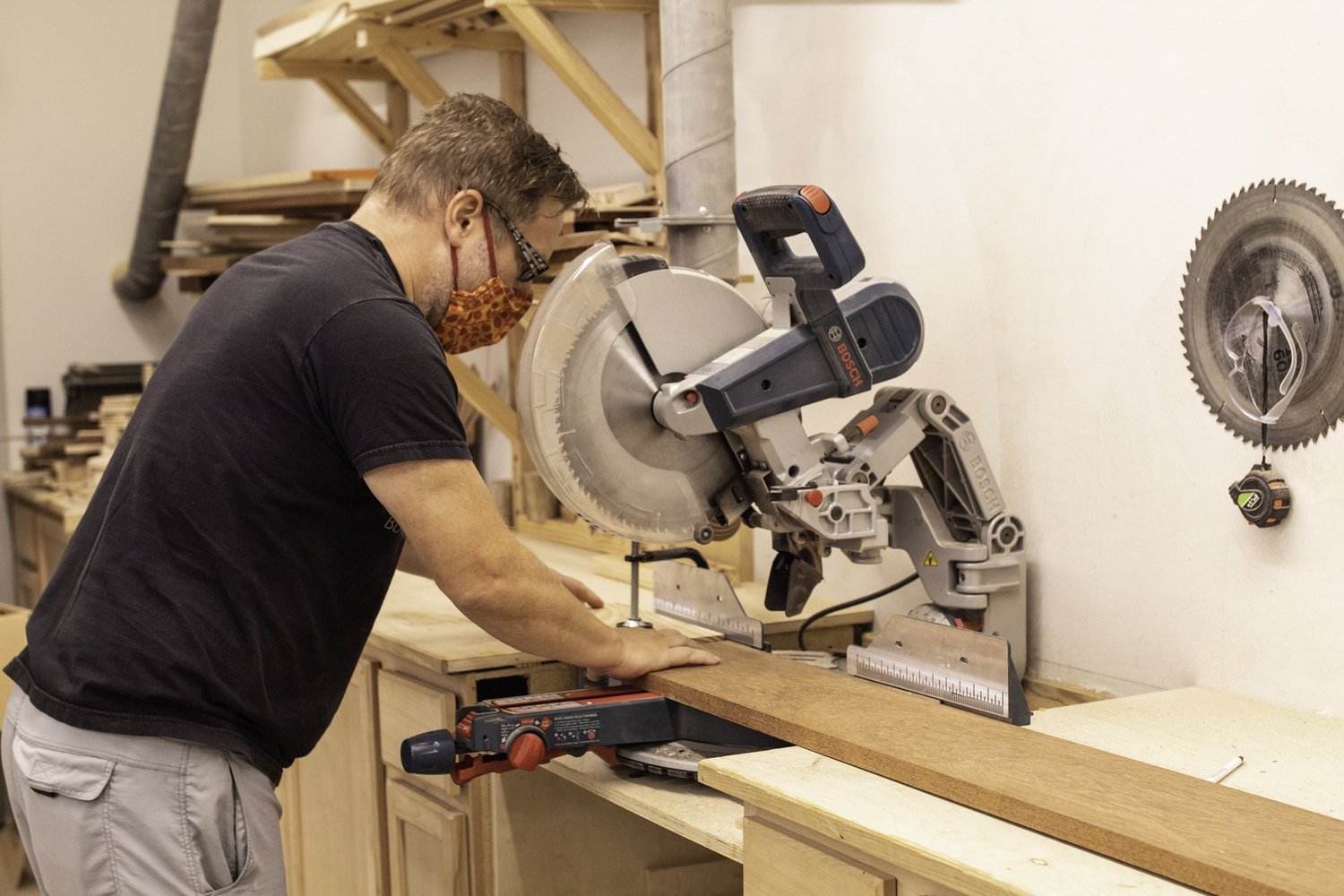 A drum maker cuts an unfinished hardwood board using a Bosch compound miter saw in a workshop.