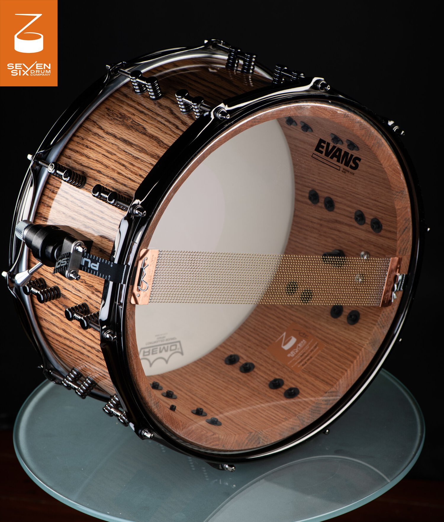 Handmade natural wood snare drum with a gloss finish, black hoops and hardware, and bronze snare wires over a clear drumhead.
