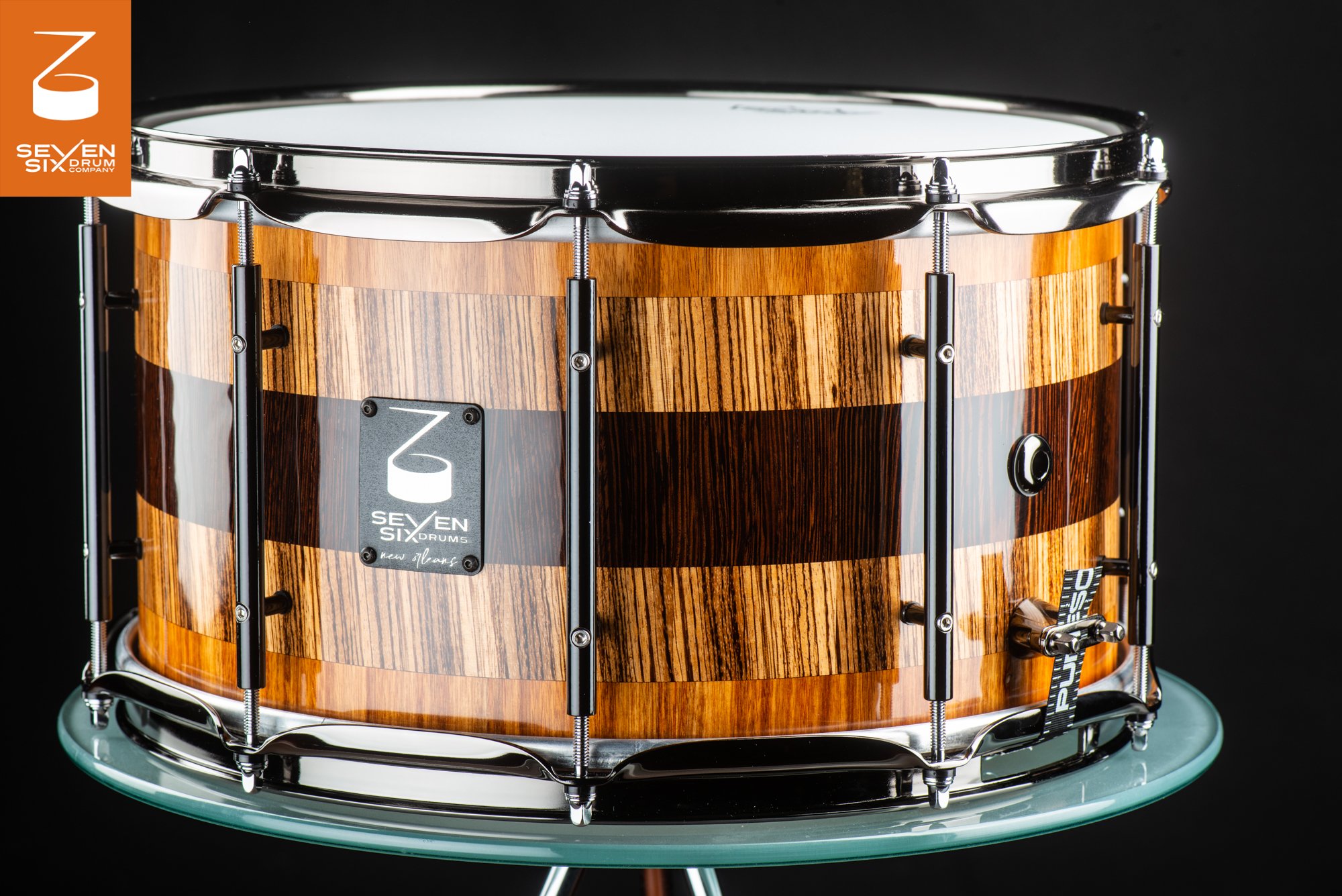 Glossy striped wood snare drum with a dark center stripe, black tube lugs, and polished chrome hoops.