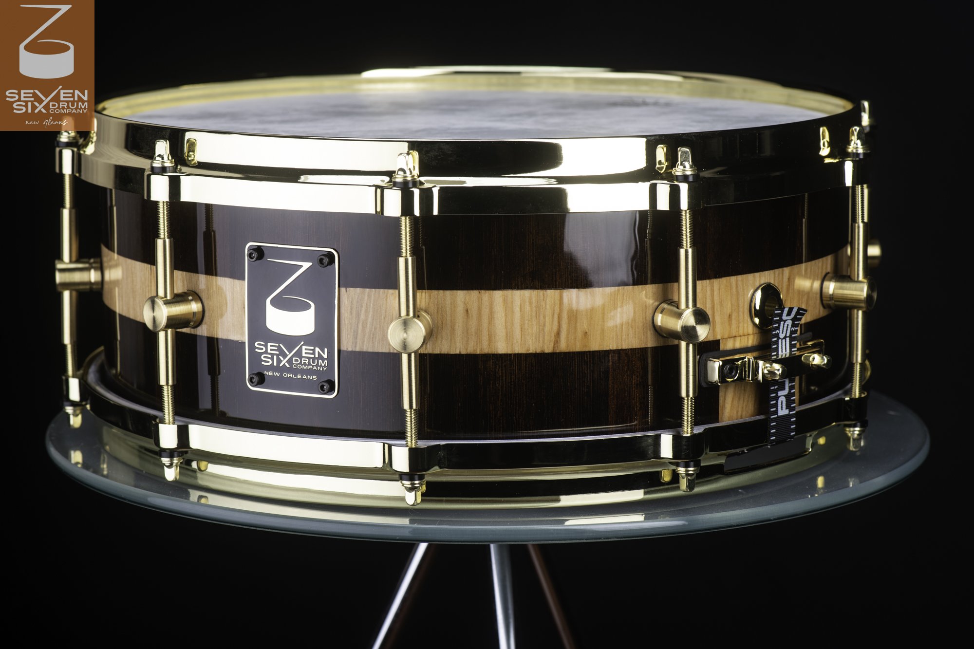 Handmade snare drum with a glossy dark and light striped wood shell, fitted with polished brass hoops and lugs.