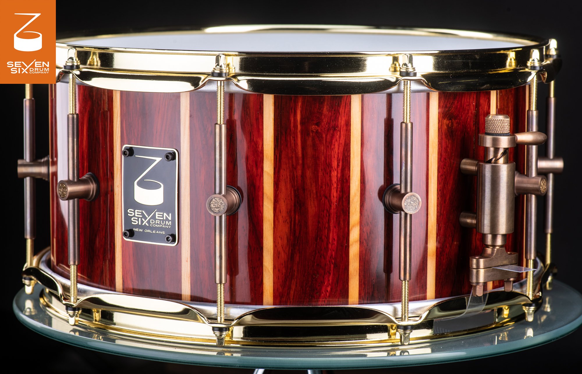 Snare drum featuring a glossy striped red and blonde wood shell, polished brass hoops, and antique bronze tube lugs and throw-off.