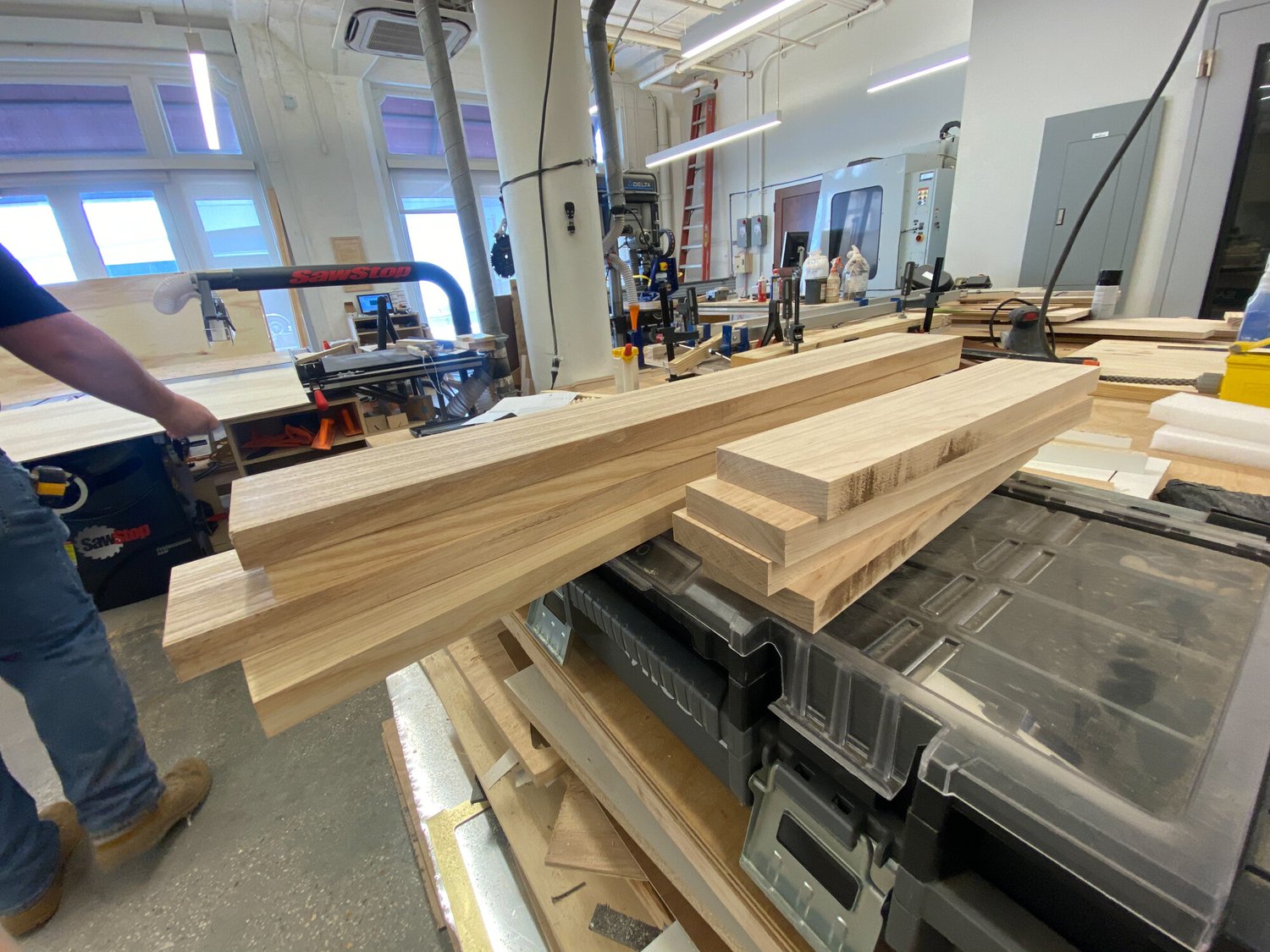 Stacks of unfinished light hardwood planks sit on a workbench in a drum shop near a SawStop table saw.
