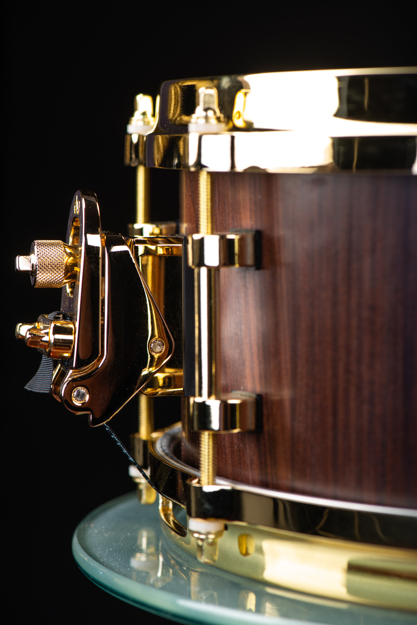 A dark wood snare drum with a satin finish, fitted with polished gold hoops, lugs, and a snare throw-off.