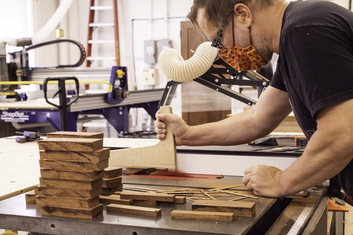 A drum maker cuts unfinished brown hardwood staves using a table saw with a wooden push stick.