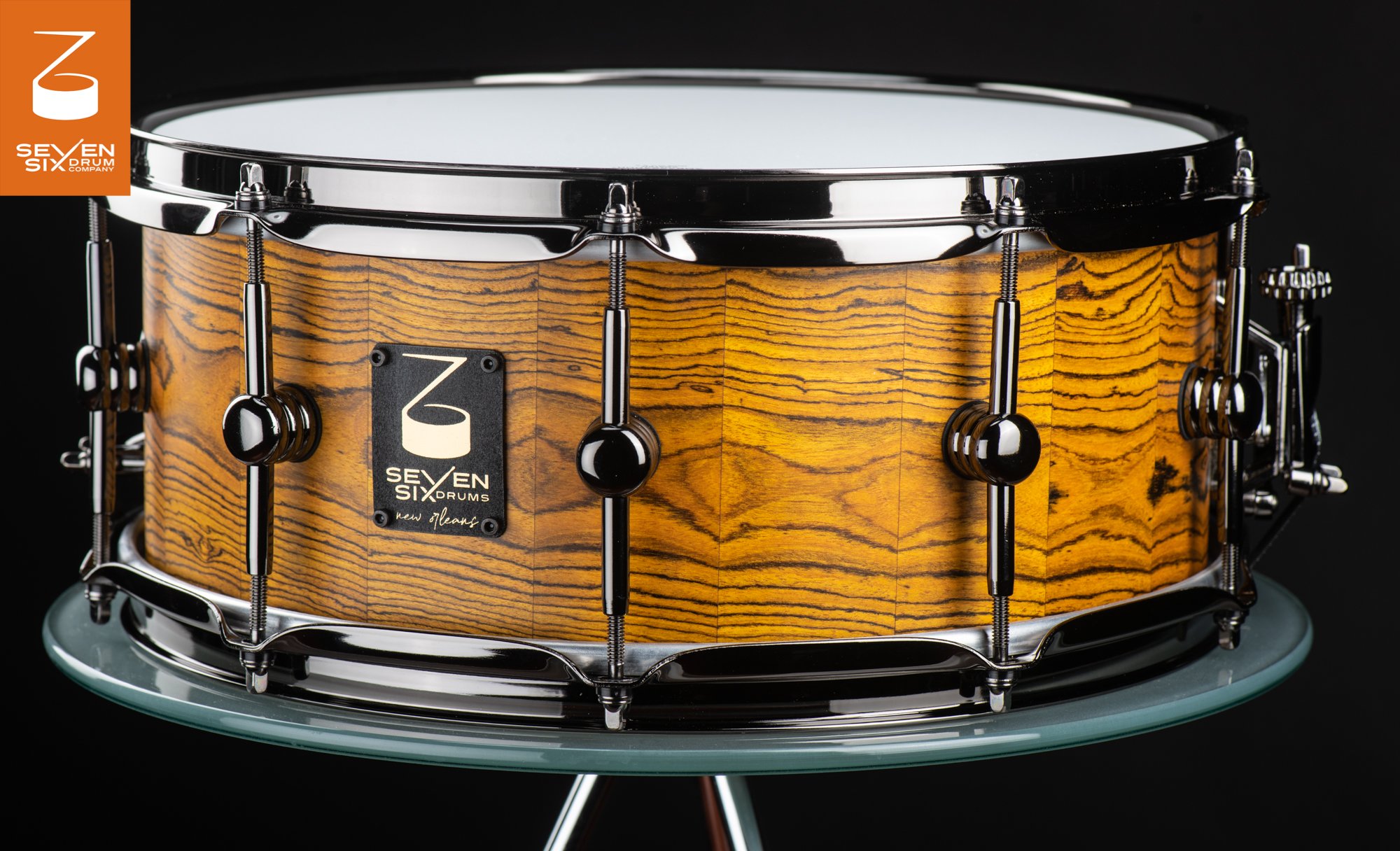 Handmade snare drum featuring a golden stave wood shell with bold grain, accented by polished black nickel hoops and lugs.