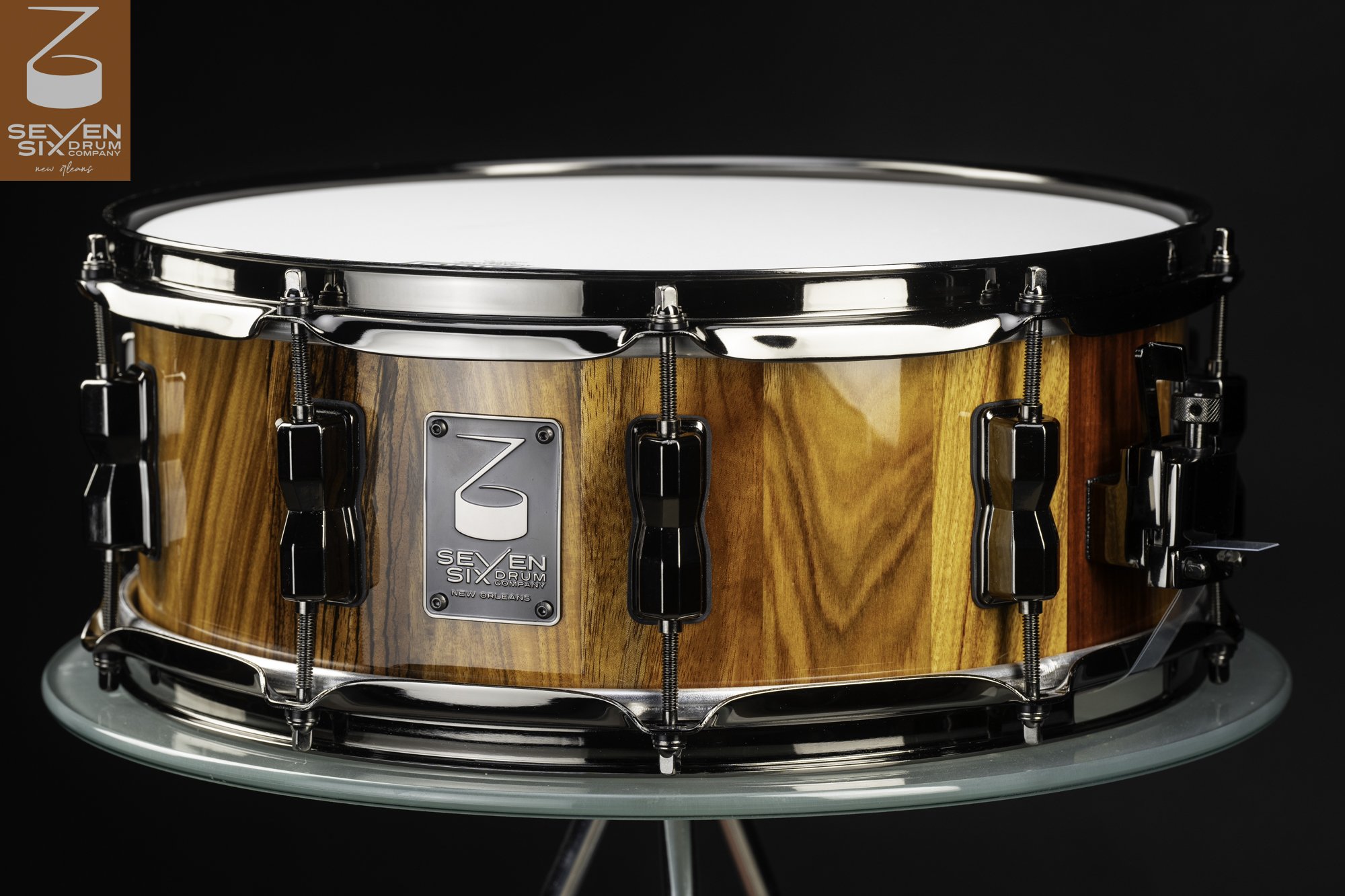 Glossy striped wood snare drum featuring black nickel hoops, lugs, and a Seven Six Drum Company badge.